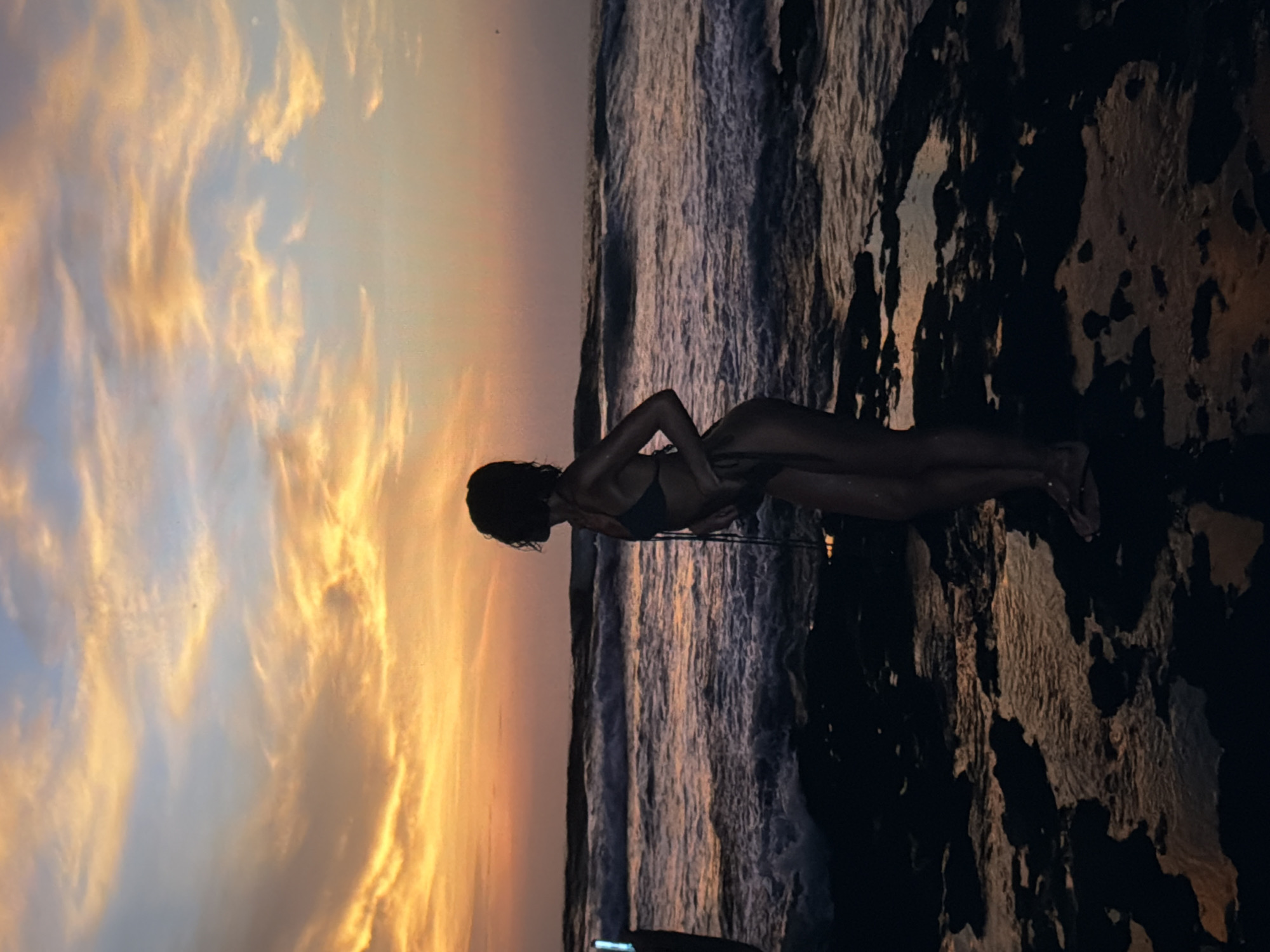 A vertical image shows a woman in a bikini lying on a rock at the shoreline, possibly at sunset, in North Kuta, Indonesia. The sky features dramatic, golden-hued clouds, while the ocean appears rough with waves. The rocky foreground is dark and textured. The woman is slender and lies facing the ocean, with her arm resting on her stomach.
