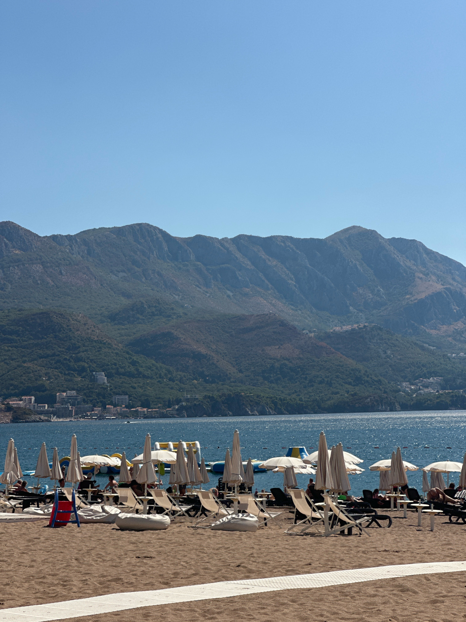 The image depicts a beach scene in Boreti, Montenegro. The foreground is dominated by a sandy beach with several rows of lounge chairs and umbrellas, some of which are occupied by people. A white wooden walkway runs horizontally across the sand. In the middle ground, the blue water of the sea is visible, with a few inflatable water attractions in the distance. The background features a lush, green mountainside dotted with buildings, and a clear blue sky above.