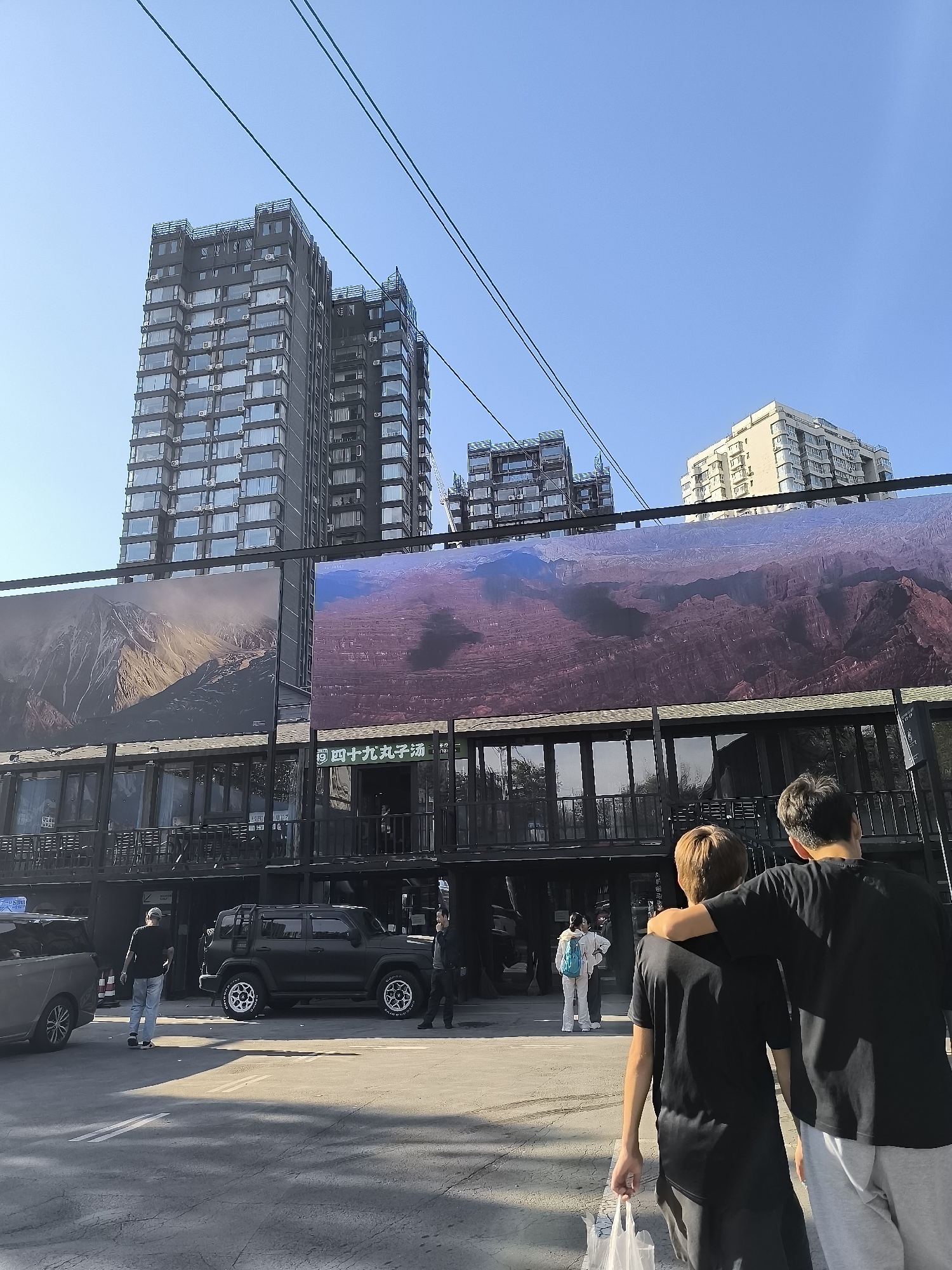 The image depicts an outdoor scene with several modern buildings in the background, including a tall, dark-colored high-rise and a lighter-colored apartment building. In the foreground, a two-story commercial building with a dark facade features large billboards displaying mountainous landscapes. Two men, dressed casually in black t-shirts and light-colored pants, are walking away from the camera, with one man's arm around the other's shoulder. In the mid-ground, several vehicles are parked, including a black SUV and a silver car. A few other individuals are visible in the distance, appearing to be pedestrians. The environment suggests an urban setting, possibly in Ürümqi, China, with clear blue skies and power lines running across the top of the frame.