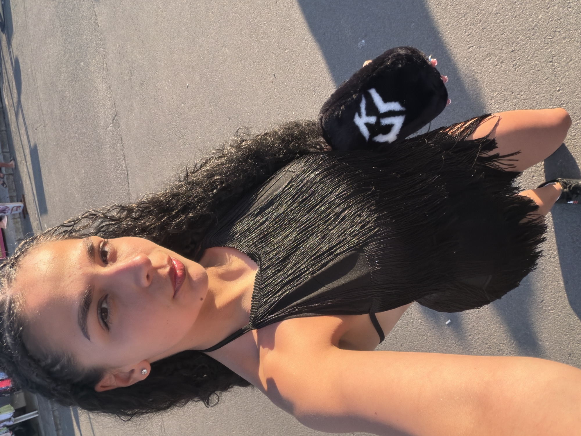 A young woman with curly dark hair stands outdoors on a paved surface, possibly a street or boardwalk. She is wearing a black dress with a fringed hem and a textured, ribbed bodice. In her left hand, she holds a small, fluffy black purse with a white emblem on it. Her right arm is extended, and she appears to be taking a selfie. Her gaze is directed towards the camera. The sunlight suggests it is daytime, likely late afternoon or early evening, given the long shadows cast on the ground. The scene is likely a public outdoor space in Nesebar, Bulgaria, as indicated by the location context. The woman's expression is neutral or slightly relaxed. There is no visible text in the image.