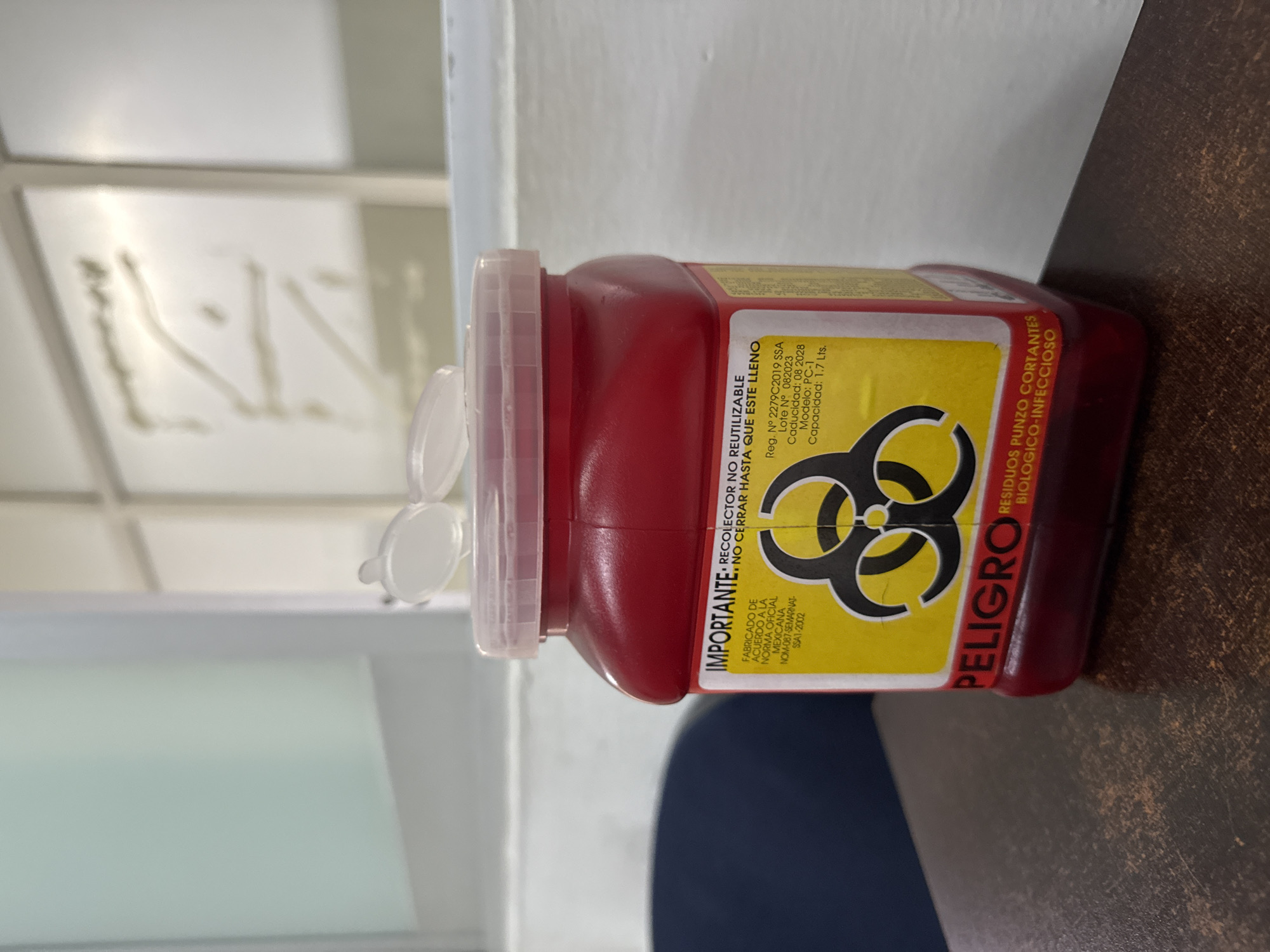 The media file displays a red biohazard sharps container, or "punzo cortantes" receptacle, positioned on a dark brown surface. The container features a translucent white, partially open cap, designed for the disposal of medical waste. A prominent yellow label is affixed to its side, clearly showing a black biohazard symbol.
The label contains text in Spanish, indicating its purpose and specifications. Key instructions read "IMPORTANTE: RECOLECTOR NO REUTILIZABLE NO CERRAR HASTA QUE ESTE LLENO" (IMPORTANT: NON-REUSABLE COLLECTOR DO NOT CLOSE UNTIL FULL). Below this, it states "FABRICADO DE ACUERDO A LA NORMA OFICIAL MEXICANA NOM-087-SEMARNAT-SSA1-2002" (MANUFACTURED ACCORDING TO MEXICAN OFFICIAL STANDARD NOM-087-SEMARNAT-SSA1-2002). Further details include "Reg. N° 2279C2019 SSA", "Lote N° 082023", "Caducidad: 08 2028" (expiration August 2028), "Modelo: PC-1", and "Capacidad: 1.7 Lts." (1.7 Liters capacity). The word "PELIGRO" (DANGER) is boldly printed vertically along one edge of the label, next to "RESIDUOS PUNZO CORTANTES BIOLOGICO-INFECCIOSO" (SHARPS WASTE BIOHAZARDOUS-INFECTIOUS).
The background consists of a light-colored wall or partition, suggesting an indoor, institutional setting, likely a medical facility in Puebla, Mexico. No specific activity is occurring, and the time of day is not discernible.