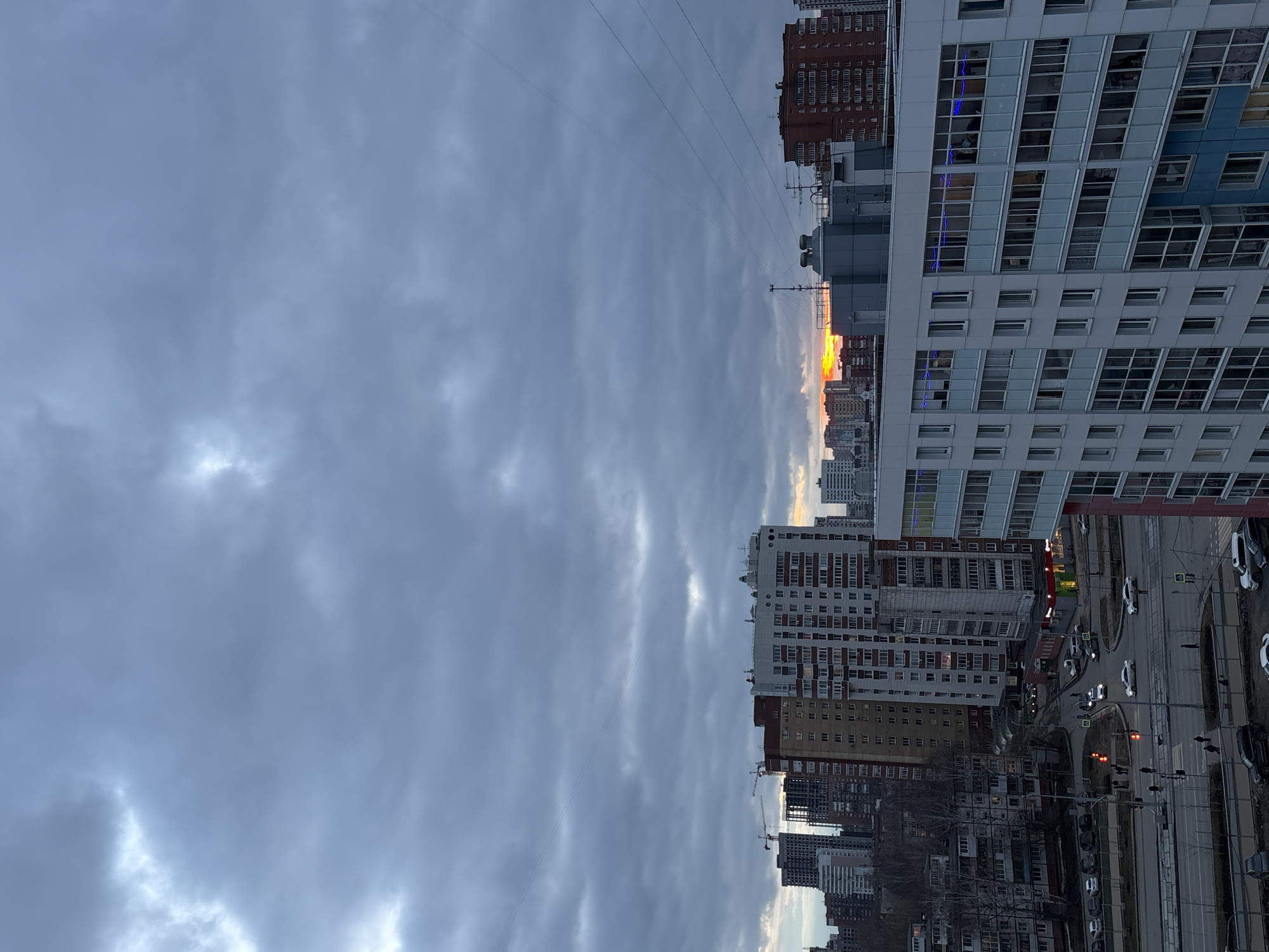 A cityscape view from an elevated position depicts a cloudy sky above several buildings and a street in Perm, Russia. The sky is overcast with gray clouds. The buildings vary in height and design, including a modern high-rise with white and blue panels, as well as older residential buildings. A street runs alongside the buildings with several parked cars. The sky suggests a setting during sunset time.