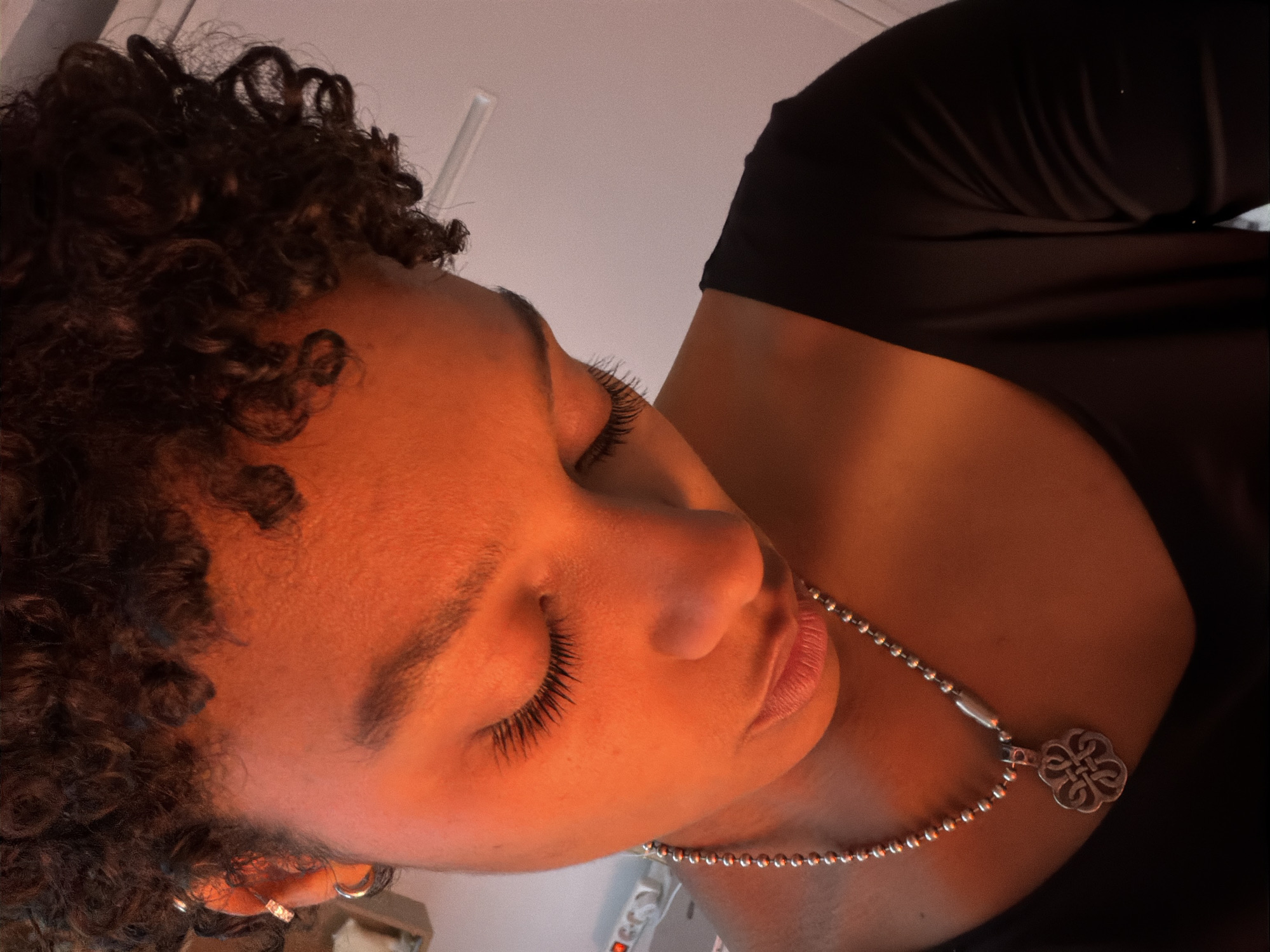 A close-up, eye-level shot depicts a person with curly brown hair, wearing a black top and a silver necklace with a clover-shaped pendant. The person's eyes are closed. The light appears warm and the background is out of focus. There are no indications of location in Paris, France.