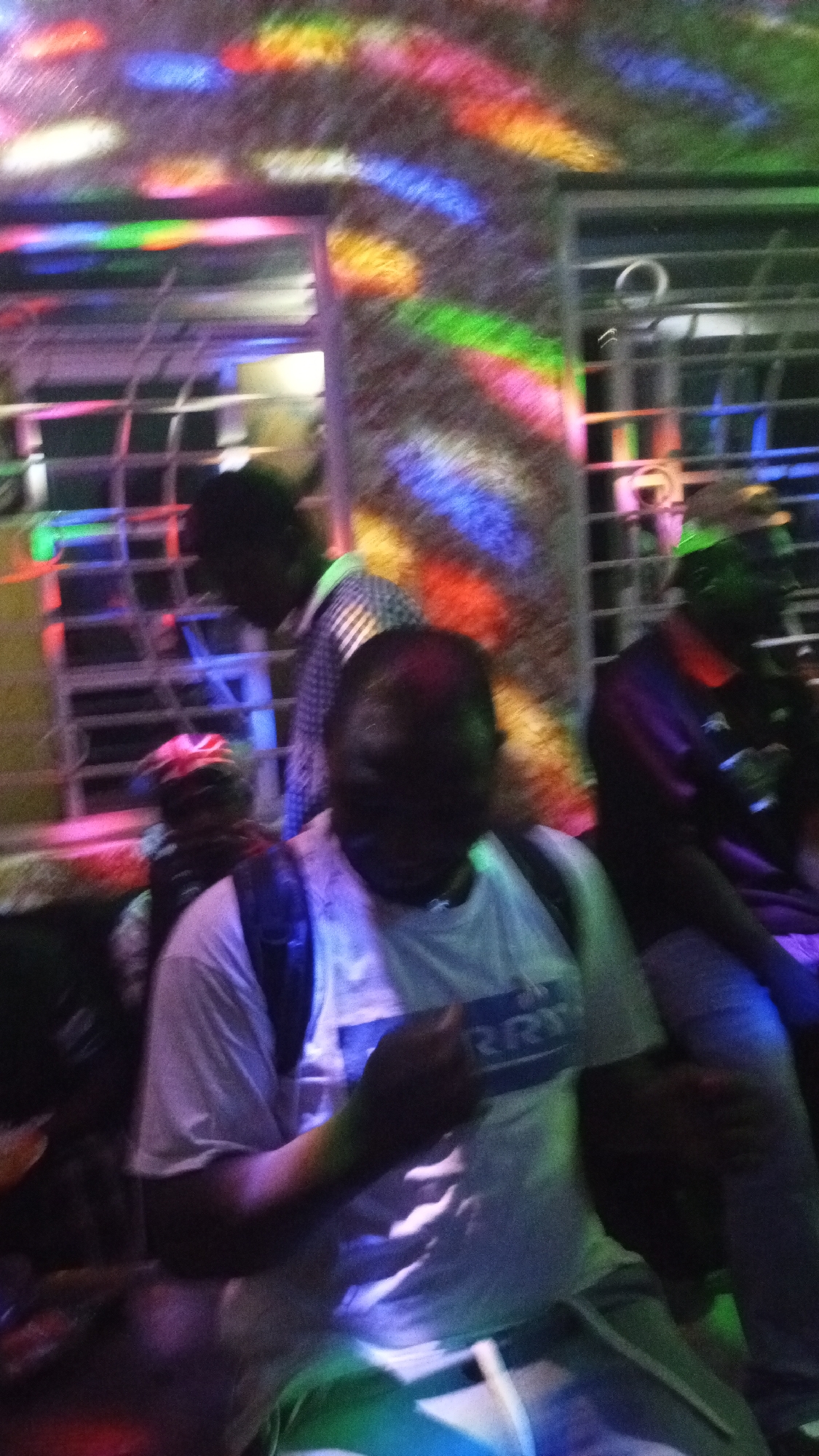 In an indoor environment illuminated by colored lights, a group of individuals is present. One person in the foreground, wearing a white t-shirt and a backpack, is partially visible. Another person, wearing a checkered shirt, stands in the mid-ground. The background shows a barred window and walls with colorful light reflections. The scene appears to be a gathering or party.