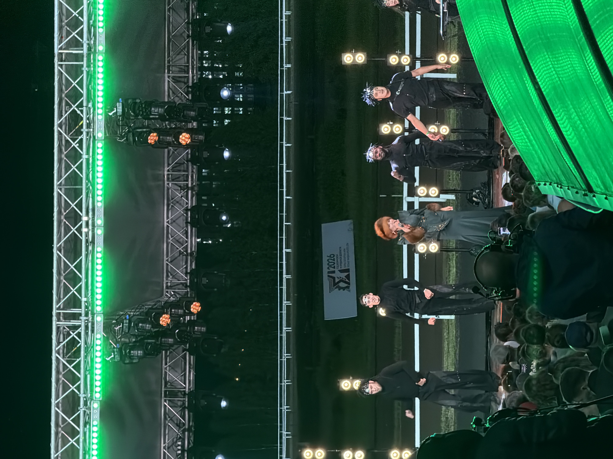 The image shows an outdoor performance stage at night, illuminated by bright green and orange lights. A group of performers, dressed in black outfits, are on stage. One female performer, wearing a furry collar and elaborate headdress, sings into a microphone. She is flanked by several male performers, some with distinct headwear, who appear to be dancing or posing. In the background, a banner with "V 2026" is visible. A crowd of spectators can be seen in the foreground, some holding cameras and phones, indicating they are recording the event. The overall atmosphere suggests a lively performance or concert.