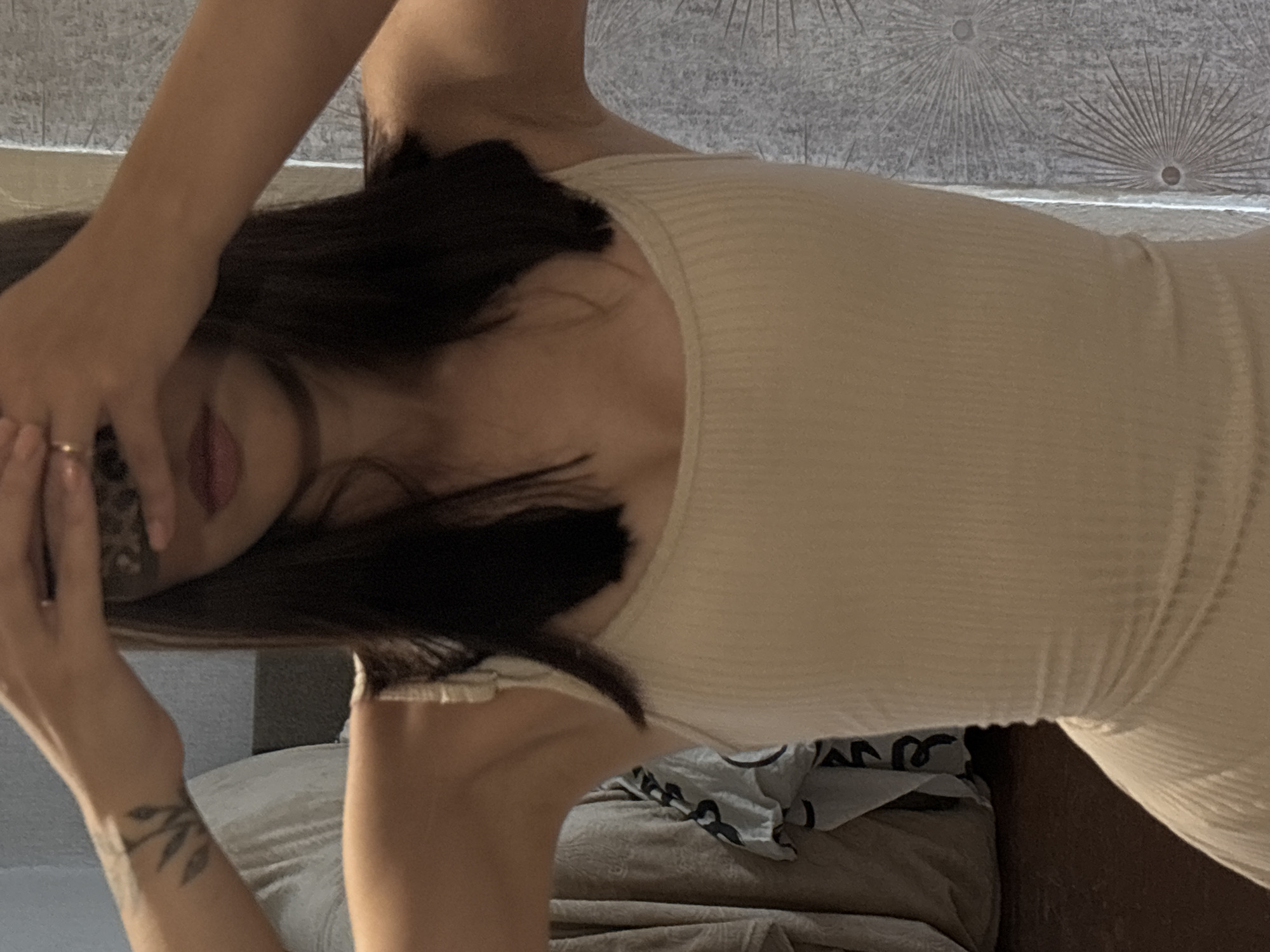 A person is captured from the chest up in a mirror selfie. They are wearing a light-colored, ribbed tank top. Their hands are raised, obscuring their face, and they are wearing a ring on their left hand. To the left of the person, a patterned fabric is visible, possibly a curtain or clothing. To the right, a wall with a textured pattern of radiating lines and circular motifs is present. The overall setting appears to be an indoor environment, likely a bedroom or dressing area. There is no indication of the specific location within the city of Inskoy, Russia.