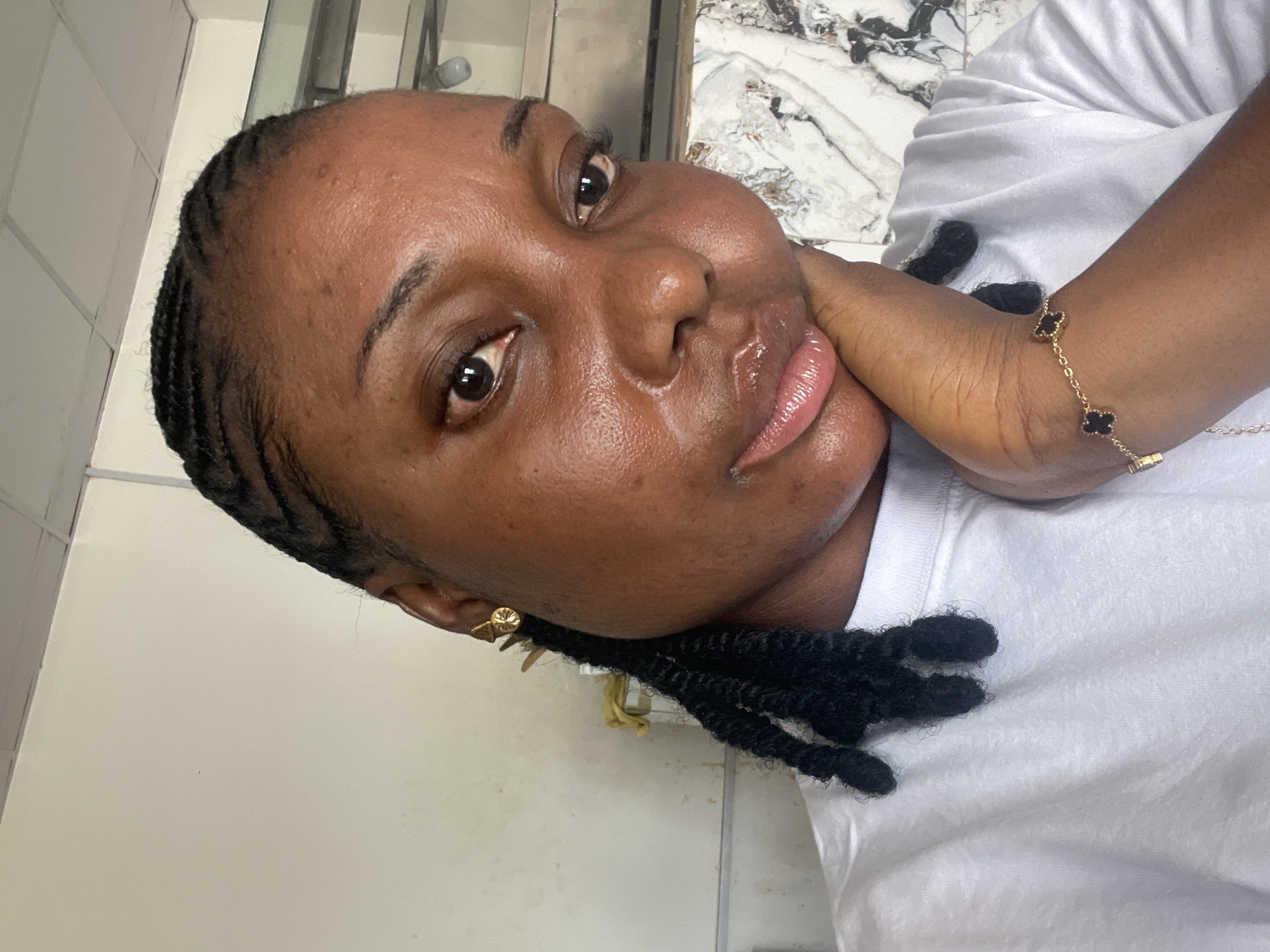 A medium close-up, tilted portrait captures a person of color, likely female, indoors in Jos, Nigeria. She looks directly at the viewer with a calm, soft expression, her face appearing moisturized and radiant. Her dark hair is styled in cornrows that transition into black, textured twists or locs. She wears a plain white short-sleeved t-shirt.
Notable details include a small gold stud earring in her visible left ear. Her right hand gently rests near her chin, revealing a delicate gold-toned bracelet on her wrist, adorned with multiple black, four-leaf clover-shaped charms.
The background is split between two surfaces: a light, plain wall or tiled surface on the left, and a white and grey marble-patterned surface on the right, possibly a countertop or decorative wall. The bright lighting suggests it is daytime. There is no visible text in the image.