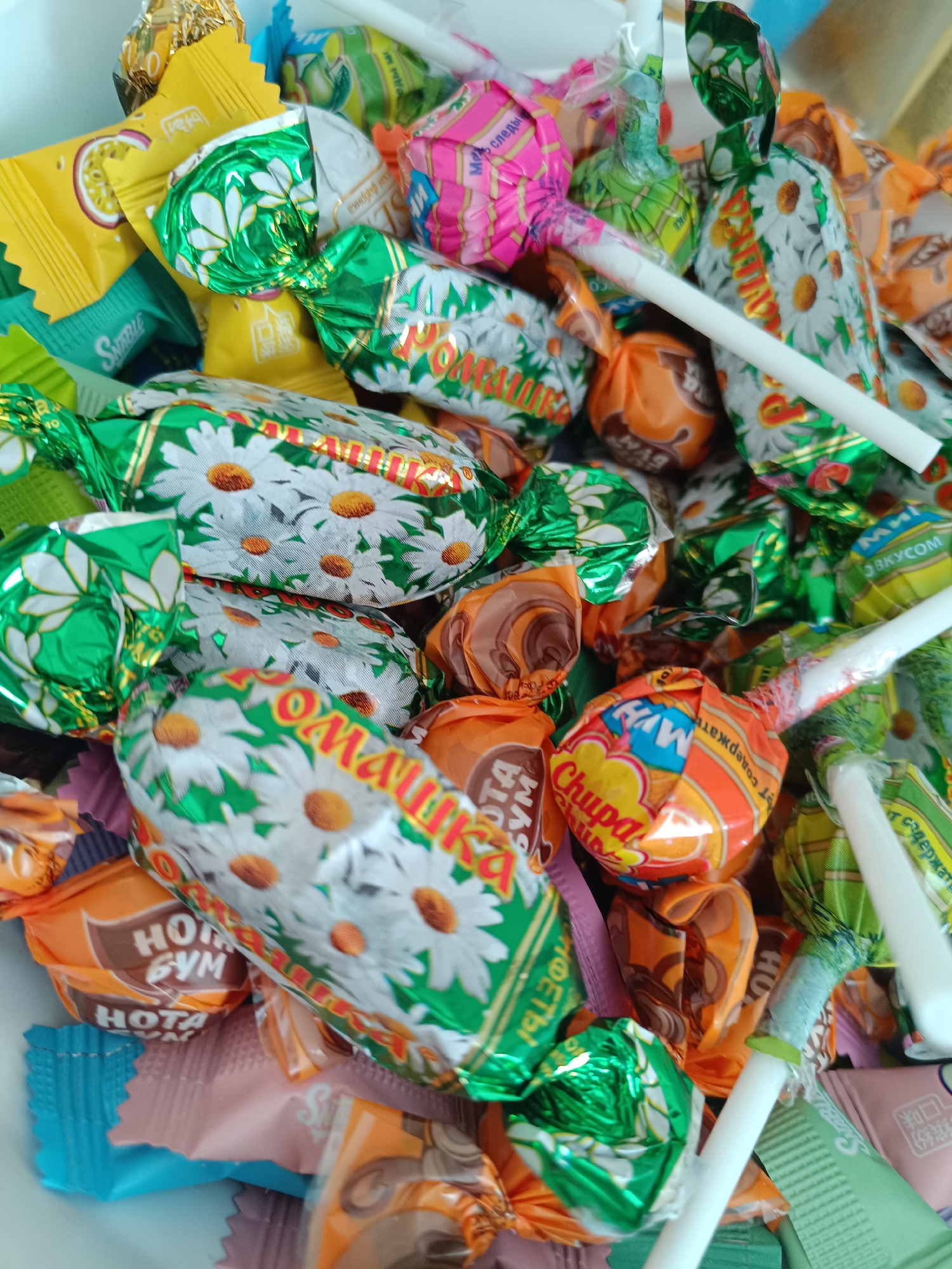 The image shows a close-up, high-angle view of a pile of assorted candies. Dominating the composition are candies wrapped in green and white foil adorned with a daisy pattern, labeled with Cyrillic text including the word "Ромашка" (Romashka, meaning chamomile). Also visible are orange and brown candies marked "Hota Gym". There are round lollipops with white sticks, some bearing "Chupa Chups" branding. Other candy wrappers in yellow, pink, and blue colors fill the remaining space, with some featuring Cyrillic and Mandarin characters. The overall arrangement suggests a casual assortment, possibly a bulk purchase or a mixed collection of treats.
There's no indication from the image where this was taken or if it's in Tobolsk. The text provides no mention of the city.