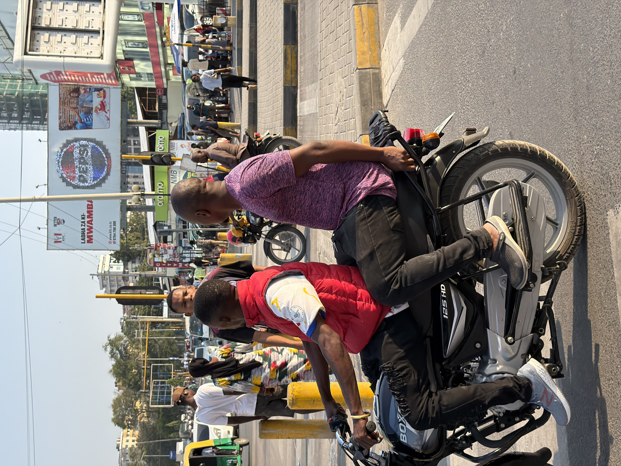 This is a photograph taken in Dar es Salaam, Tanzania, featuring a busy urban street scene. The foreground is dominated by two men riding a motorcycle. The driver, wearing a red puffer vest over a white t-shirt with a colorful abstract pattern, is positioned in the front. He has dark skin, short black hair, and appears to be looking forward. The passenger, on the back, is wearing a purple, heathered t-shirt and dark pants. He has dark skin and short hair, and is looking to his left. The motorcycle is black with silver accents, and the model "BOX 125 HD" is visible on its side.
In the background, the street is filled with various vehicles, including cars, a green tuk-tuk on the left, and other motorcycles. Pedestrians are also present, walking along the sidewalks and crossing the street. Several large billboards are visible in the distance, including one advertising Pepsi with a circular logo and images of people enjoying drinks, and another for KFC. Traffic lights and street lamps are also present, indicating an intersection. Buildings of varying heights, some with scaffolding suggesting ongoing construction, line the street. The sky is clear and blue, suggesting a sunny day. The overall atmosphere is one of a bustling city environment with significant traffic and commercial activity.
