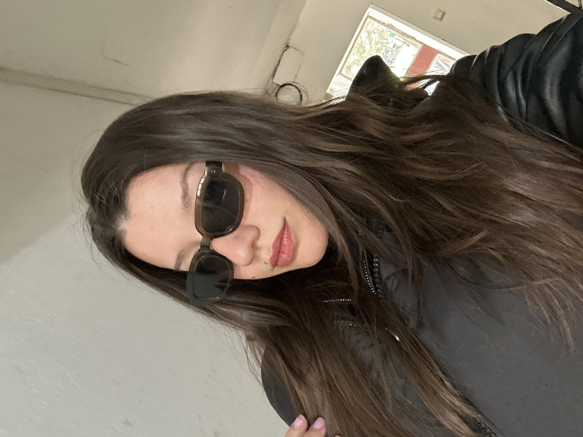 A low-angle close-up portrait depicts a young woman with long, brown, wavy hair wearing dark sunglasses. She is positioned indoors, near a window visible in the background. The woman is wearing a black, possibly leather, jacket. The indoor environment appears to be a room with a white ceiling and wall. The location is specified as Burgas, Bulgaria.