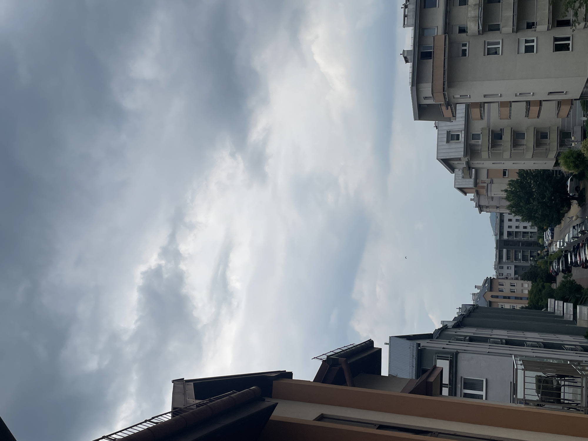 The image shows a view of apartment buildings and a street with parked cars under a cloudy sky. The buildings are multi-story structures with balconies and windows. On the left side of the image, a building with orange and brown exterior is partially visible. A street lined with parked cars runs through the middle ground and background, with trees and other buildings visible beyond. The sky is overcast with gray and white clouds. There is a small bird in flight in the upper portion of the image. The scene appears to be an urban residential area.