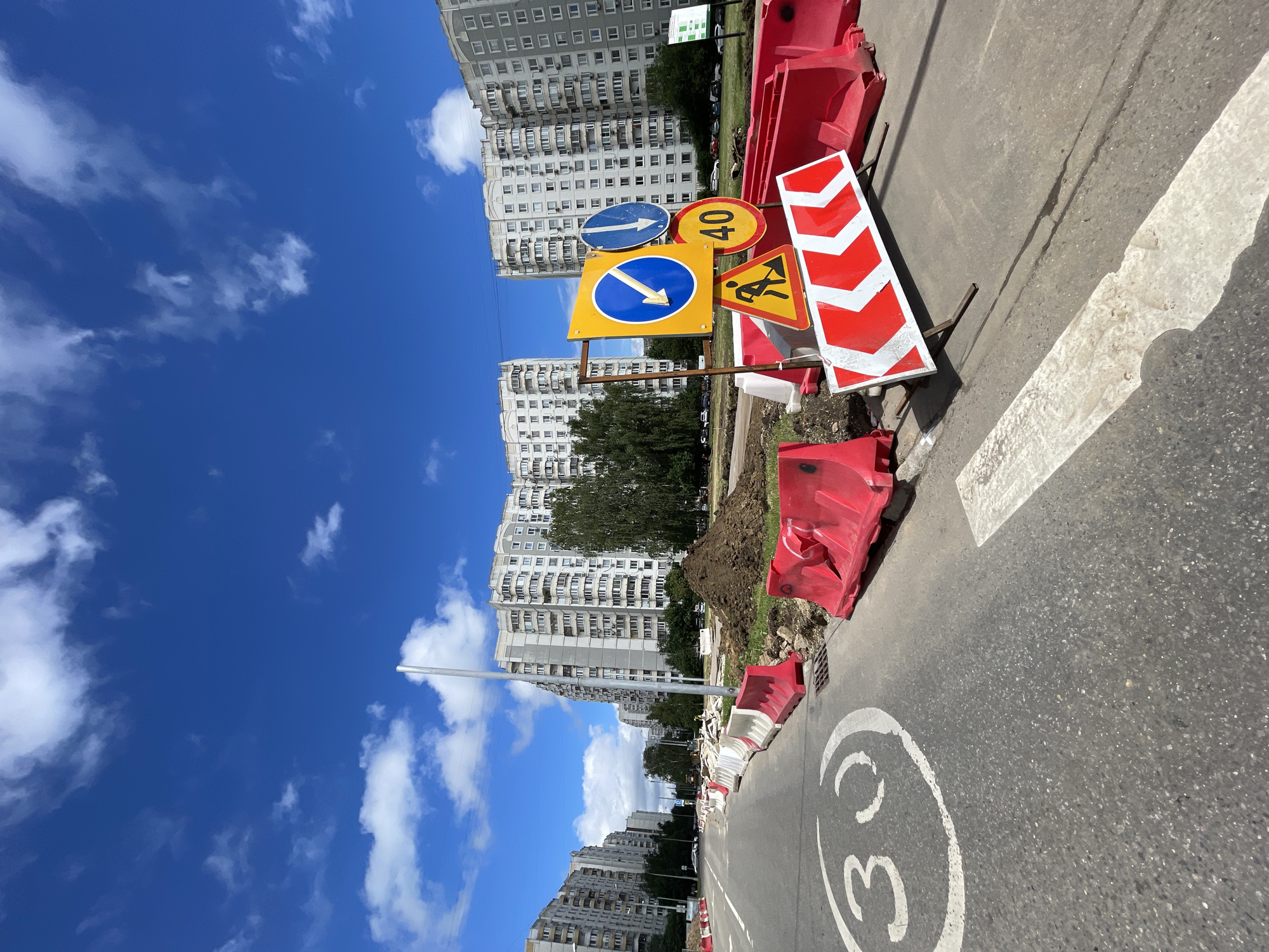 This is a ground-level, wide-angle photograph of a street undergoing construction in Moscow, Russia. The scene is dominated by large, multi-story apartment buildings, characteristic of Moscow's urban landscape. In the foreground, the asphalt road features a painted "30" speed limit marking and a white pedestrian crossing. Construction barriers, made of red and white plastic, line the road, indicating a work zone. Several traffic signs are visible, including a mandatory direction sign with a left arrow, a blue circular sign with a downward arrow, a circular yellow sign with the number "40," and a triangular yellow sign depicting a worker. A large pile of dirt is situated near the barriers. The sky is a vibrant blue with scattered white clouds. The overall impression is one of an active construction site within a residential area.
