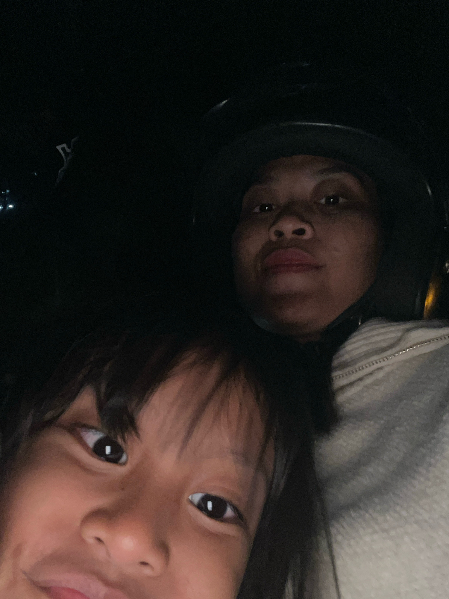 This is a close-up, selfie-style photograph taken at night, likely in Kecamatan Pati, Indonesia. Two individuals are visible: a child in the foreground and an adult in the background. The child is positioned close to the camera, filling the lower half of the frame with their face turned slightly to the left. They have dark hair, dark eyes, and appear to be smiling. The adult is behind the child, wearing a dark helmet, and their face is partially visible from the eyes upwards. They are wearing a light-colored, zippered jacket. The overall lighting suggests the photograph was taken in a low-light environment, possibly outdoors on a street or during travel, as indicated by the helmet and the darkness of the surroundings. The quality of the image is somewhat grainy, consistent with a night-time photo taken with a mobile device.