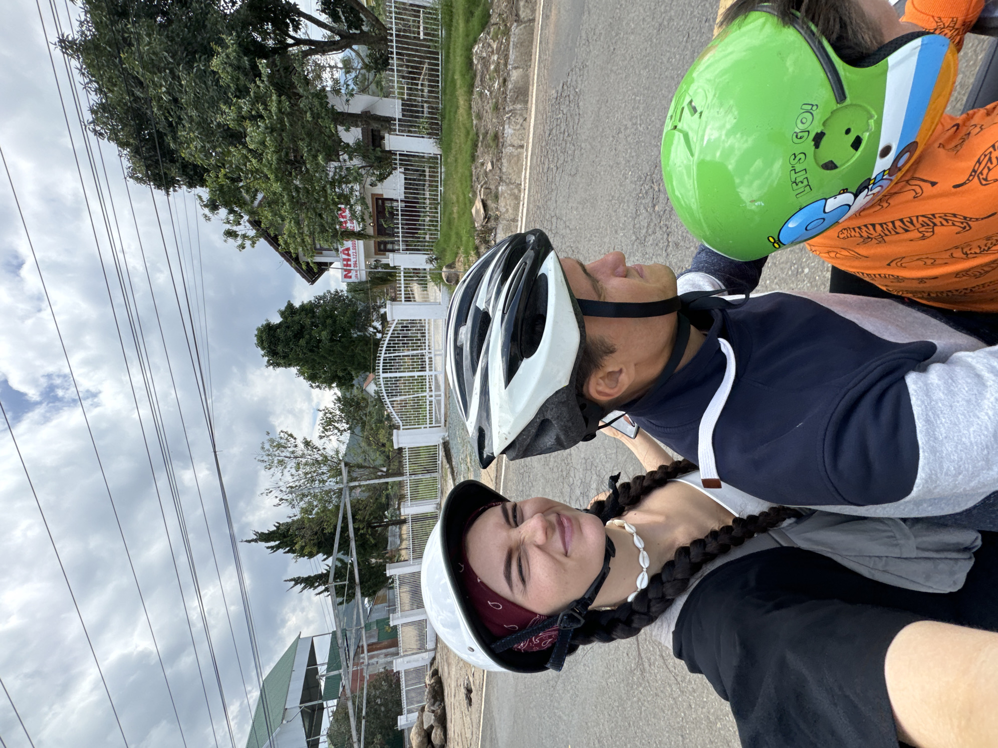 The image captures three people wearing helmets, likely riding a motorbike on a paved road. The person in the foreground wears a white helmet with a red bandana underneath and has braids. A man with a black and white helmet is behind them. A child with a green helmet is seated behind the man. The backdrop features buildings and lush greenery, typical of Thôn 5, Vietnam. Utility poles and wires are visible against a cloudy sky. The road surface shows signs of wear. The image appears to be a selfie or candid shot taken during a motorbike ride.