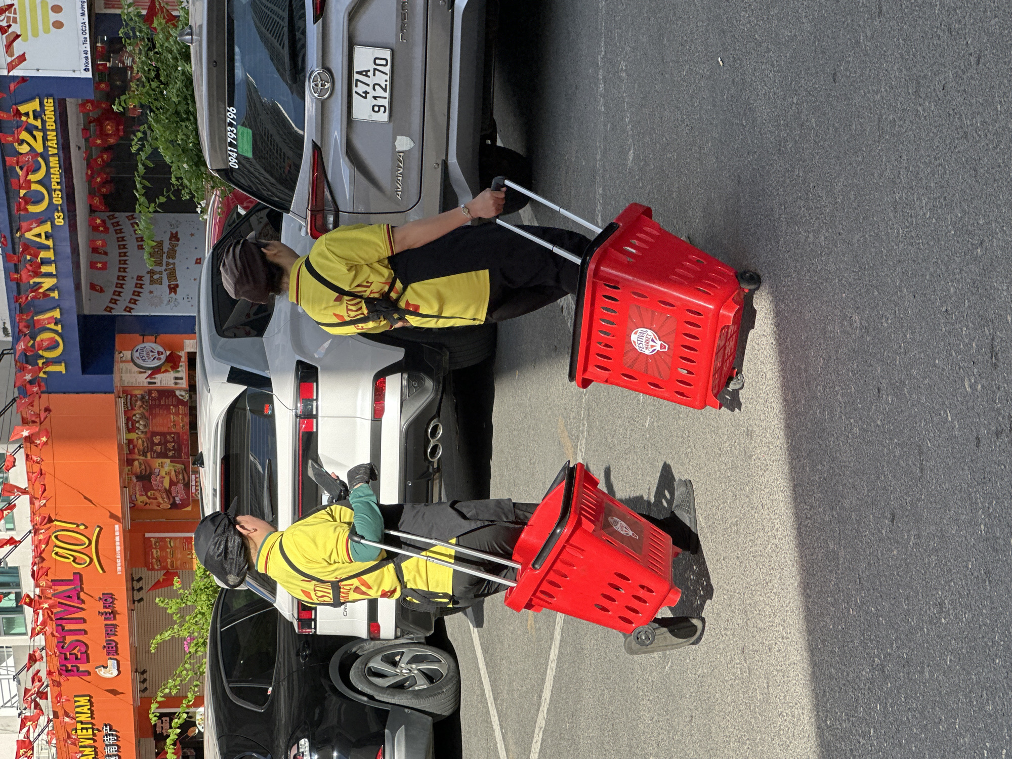 Two individuals, dressed in yellow shirts with black suspenders and black pants, are walking on a paved surface in what appears to be a parking lot. The person on the left is wearing a black cap, and both individuals are pulling bright red rolling shopping baskets. The basket on the left has a logo that reads "FESTIVAL MARKET". Behind them, several cars are parked, including a silver Toyota Avanza with a license plate ending in "70". The background features buildings adorned with red flags and banners, with signage that includes text in Vietnamese, such as "FESTIVAL GO!" and "TOA NHA OC2A". The environment suggests an urban setting in Bac Nha Trang Ward, Vietnam, possibly during a festive event. The lighting indicates it is daytime, with strong shadows cast on the ground.