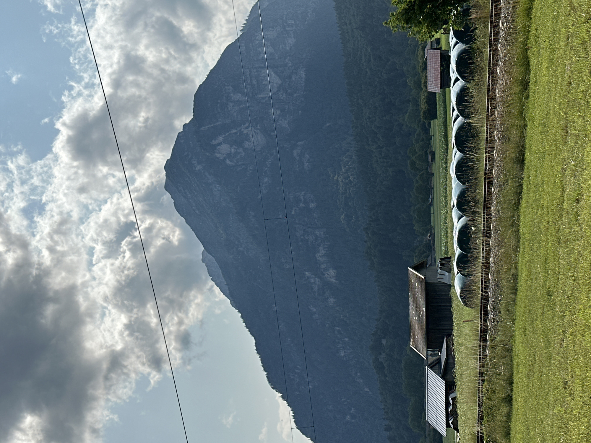 The image shows a rural landscape with a large mountain in the background. In the foreground, there are two farm buildings, one with a metal roof and another with a darker, sloping roof. In front of these buildings, there is a row of large, round hay bales wrapped in green plastic, arranged along a set of train tracks. Beyond the hay bales, there is a field of tall grass. The mountain in the background is covered in trees at its lower slopes and shows rocky, bare terrain higher up. The sky is partly cloudy with sunlight breaking through. The scene suggests a pastoral setting in Farchant, Germany, likely during the daytime under a partly cloudy sky. There are no people or animals visible in the image. The overall impression is one of a serene, agricultural environment dominated by the presence of the imposing mountain.