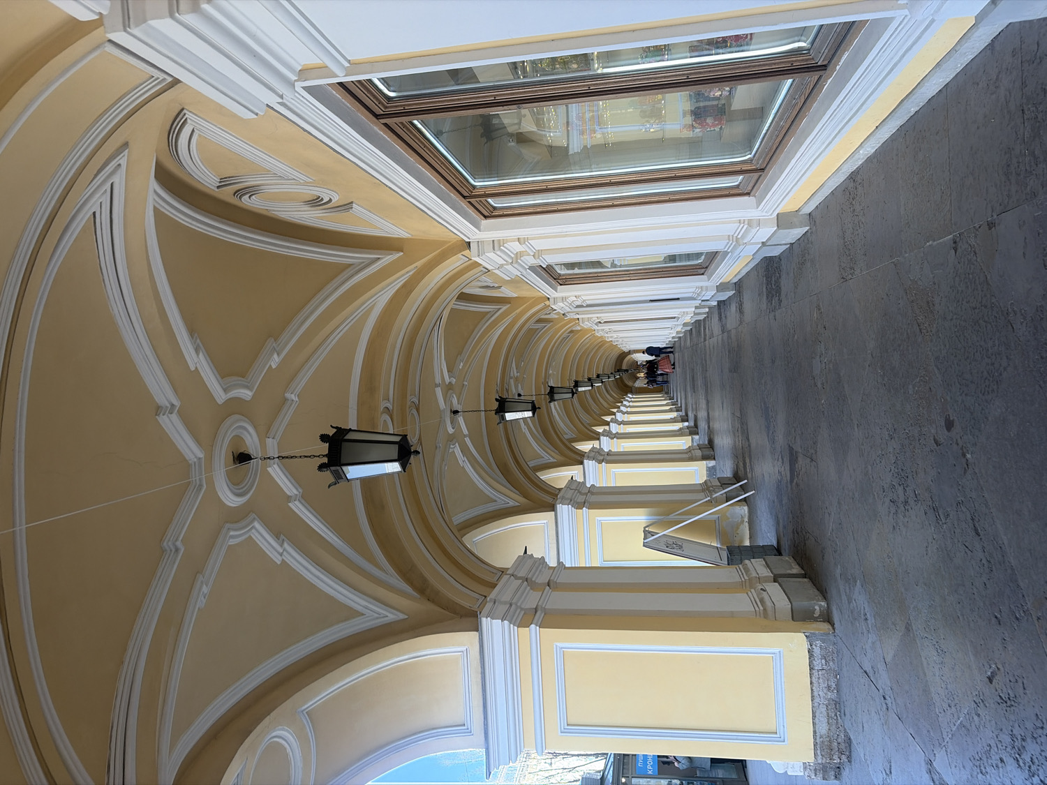 The image captures a long, ornate covered walkway or arcade, characterized by repeating architectural elements. The perspective draws the eye down the length of the passage, which features a series of arches and a vaulted ceiling, all painted in a warm light yellow or cream color with decorative white trim. Hanging lanterns with dark frames are suspended at regular intervals from the ceiling, adding to the classical aesthetic.
To the right, a continuous line of shop windows or display cases extends along the walkway, reflecting the interior and some indistinct shapes. The floor is a dark, textured paved surface, suggesting stone or concrete. In the far distance, several indistinct human figures can be seen walking, indicating that this is a public thoroughfare or commercial space.
The lighting is soft and even, suggesting either an overcast day or an interior setting with indirect illumination, possibly from skylights or the open ends of the arcade. There are no harsh shadows, giving the scene a serene and well-lit appearance. The architectural style, with its classical arches and detailed trim, suggests a historical building.
Visible text on a small sign attached to a pillar on the lower left edge includes partial Russian words, such as "...KPOH..." and "ПУТЕШЕСТВИЕ" (JOURNEY), which, combined with the architectural style, strongly points to a location in Russia, likely Saint Petersburg, as hinted. The overall atmosphere is one of elegant grandeur and quiet activity.