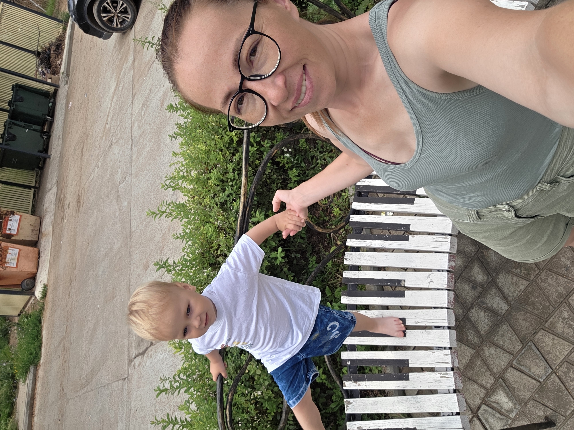 A woman with glasses and a green tank top is taking a selfie with a young boy standing on a piano-key-themed bench. The boy is wearing a white t-shirt and blue denim shorts. The woman's right hand is extended towards the boy, who is reaching out with his left hand. The background shows a paved area, green bushes, and some trash bins in the distance. The scene appears to be outdoors in a residential or public area. The woman's glasses are black-rimmed and round. The bench is made of wood, painted with black and white keys. The paving stones are irregularly shaped and laid in a pattern. The overall lighting suggests it is daytime.
