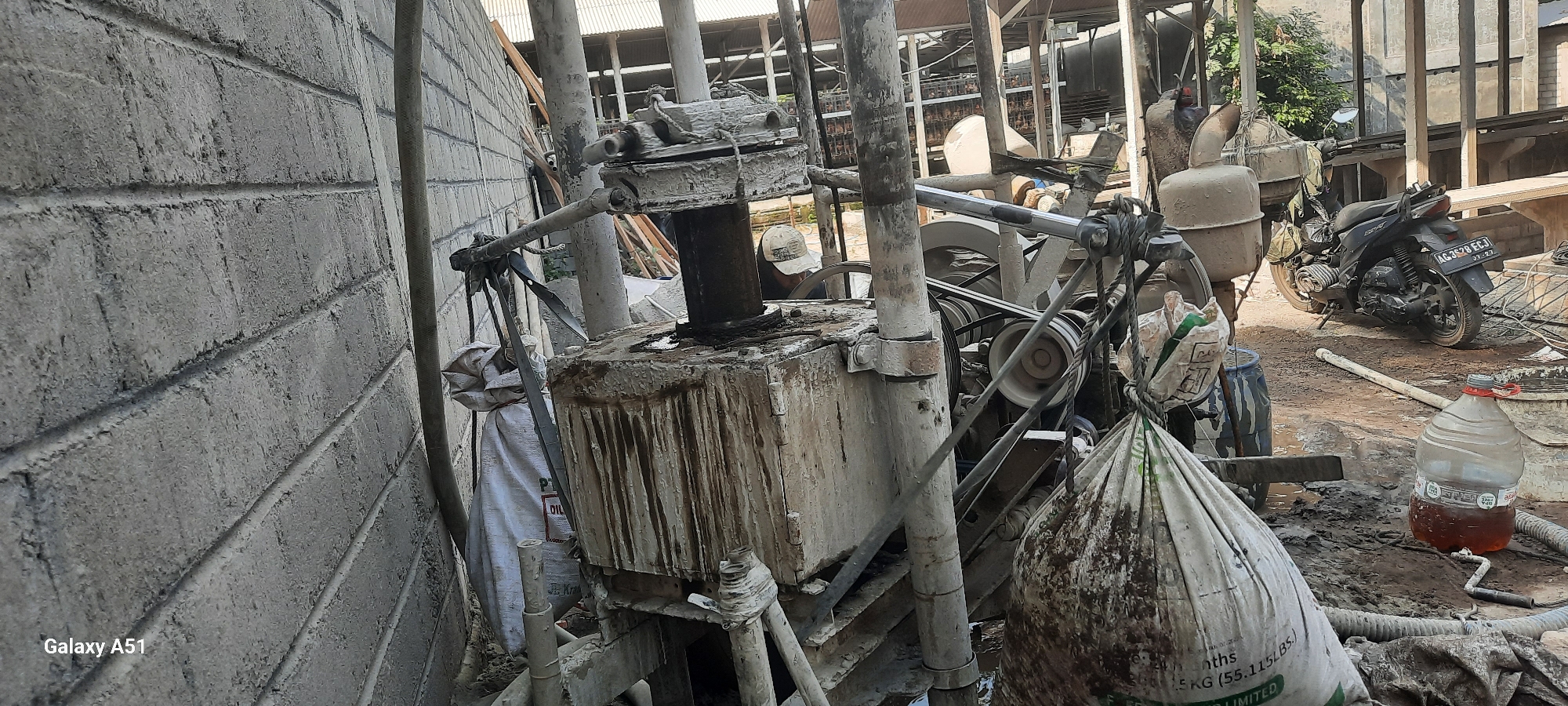 In Ploso, Indonesia, a construction site is depicted, focusing on drilling equipment. A large concrete wall runs along the left side of the frame. In the center, a drilling machine is partially obscured by a large, dirt-covered bag hanging from a strap. The machine itself appears weathered and dirty, with exposed gears and pipes. A worker, wearing a cap, is visible in the background, seemingly operating or attending to the machinery. To the right, a black motorcycle with the license plate "AG 3628 ECJ" is parked. A large plastic bottle filled with a brown liquid stands on the muddy ground, along with various construction materials and debris. The overall environment is muddy and appears to be an outdoor industrial or construction area.