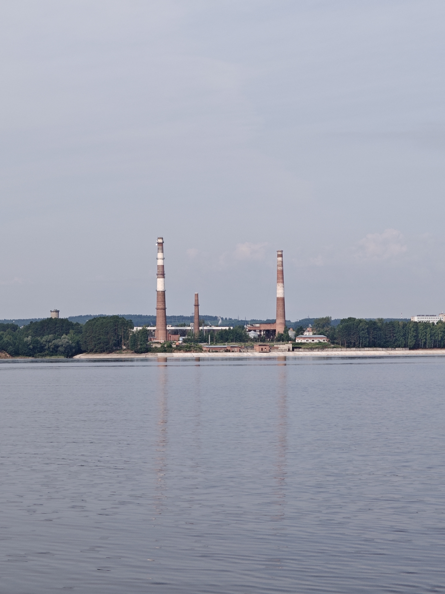 The image captures a wide view of the Kama River in Chaykovsky, Russia, with an industrial complex situated on the opposite bank. The foreground is dominated by the calm, grey-blue water of the river, reflecting the overcast sky and the structures on the shore.
On the far bank, a notable industrial facility stands, characterized by several tall, brick chimneys. The most prominent of these chimneys is striped with alternating bands of red brick and white. To the left, a shorter, similarly striped chimney is visible. Behind the chimneys, various industrial buildings with red roofs can be seen, nestled amongst a dense line of green trees, primarily pine and deciduous varieties. A water tower, cylindrical and light-colored, rises above the treeline on the far left. Further to the right, beyond the industrial buildings and trees, a cluster of modern, multi-story white buildings suggests a residential or administrative area.
The overall atmosphere is one of quiet industrial activity on a cloudy day. The scene is static, with no visible people or ongoing actions. The environment is characterized by the juxtaposition of the natural river and forest with the man-made industrial structures. The lighting is diffused due to the overcast conditions.
