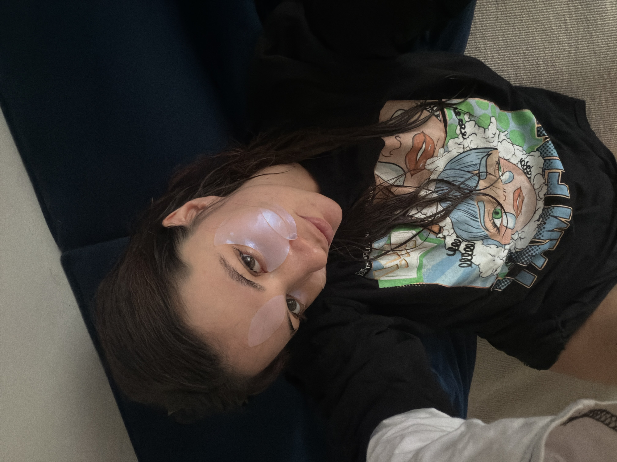 A woman is lying on a dark blue sofa. She has fair skin, dark hair and is wearing eye patches. She is wearing a black t-shirt with a large cartoon graphic. The location is unknown and there is no indication that it is in Belgrade, Serbia. The woman is interacting with the camera by looking directly into it. The environment appears to be indoors.