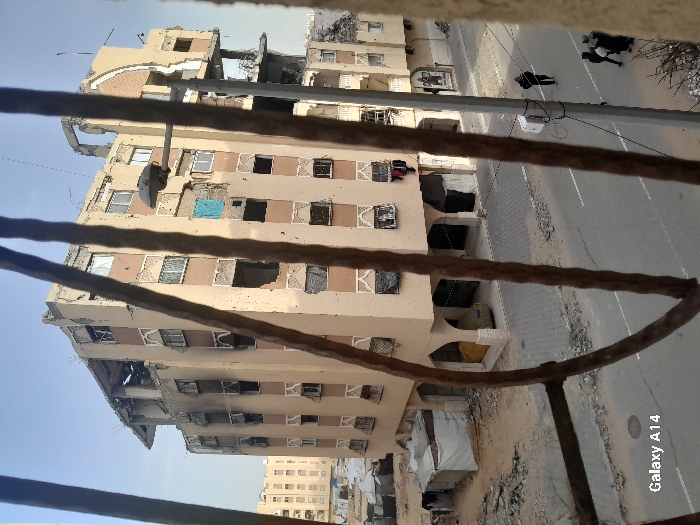 This image, captured from an elevated perspective, likely a window, frames a damaged urban streetscape in Khan Yunis, Palestinian Territory, through foreground vertical metal bars.
The central subject is a heavily damaged multi-story building, appearing to be approximately seven stories tall, constructed of light-colored (beige/tan) concrete or stucco. Its uppermost floor and roof show significant structural collapse with exposed rebar. Multiple windows throughout the building are either shattered, missing, or covered with dark tarpaulins or blankets. Dark soot or smoke damage is evident around many window openings, particularly on the upper-left side of the facade, suggesting past fires. A bright blue fabric hangs from a window on an upper-middle floor. The lower levels feature arched entryways, some partially obscured or damaged. On the far right, a section of the building appears to have sustained severe vertical damage or partial collapse.
To the immediate left of the main building, an area of disturbed earth and rubble leads to several small, makeshift shelters constructed from corrugated metal and white tarpaulins. In the far left background, another multi-story building is visible, appearing less damaged.
A paved asphalt road with faded white markings runs across the mid-ground. Debris, including concrete fragments and dirt, is scattered on and alongside the road. A street light pole with a luminaire and visible wiring stands on the right side of the road.
Several individuals are present on the street: one person in dark clothing walks alone away from the camera on the right side of the road, and further down, a group of three individuals, including at least one child, also in dark clothing, walks in the same direction. The sky is clear and light blue, indicating daylight. The bottom-left corner of the image contains the text "Galaxy A14".