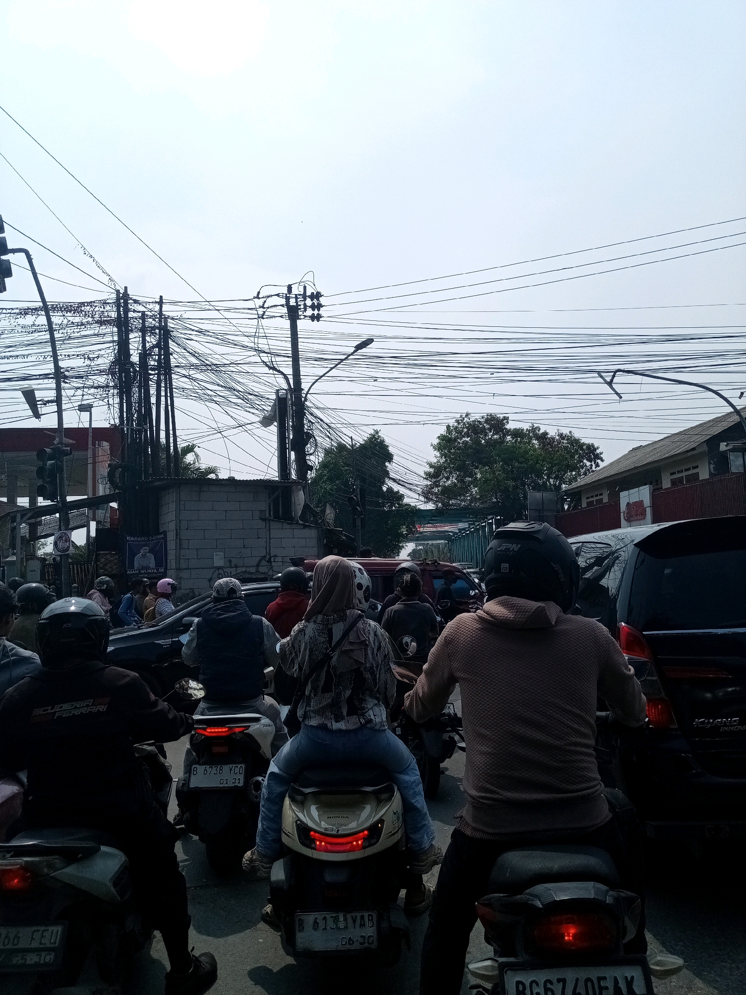 This is an outdoor, eye-level shot of a congested traffic scene in Karangsatria, Indonesia, likely taken during daytime given the bright sky. The foreground is dominated by the rear ends of several motorcycles and their riders, indicating a slow-moving or stopped traffic situation. The riders are wearing helmets, and some are wearing casual clothing, including a woman with a patterned hijab and a man in a hooded sweater.
The middle ground shows more motorcycles and the sides of cars, suggesting a multi-lane road. A notable detail in this area is a poster on a brick building, with text visible that includes "KARANG SATRIA". This confirms the location as Karangsatria. The building itself appears to be a small, functional structure, possibly a shop or maintenance area.
The background is characterized by an overwhelming network of electrical wires strung between numerous poles. These wires create a dense, tangled canopy overhead, a common sight in many urban and semi-urban areas in Indonesia. Beyond the wires, buildings with red roofs and trees are visible, suggesting a developed urban environment. The sky is overcast and bright, with a diffused sunlight. The overall impression is one of everyday urban life, with the traffic and overhead infrastructure being prominent elements of the scene.