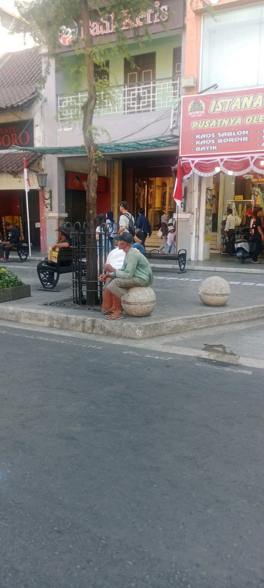 The image depicts a street scene in the city of Yogyakarta, Indonesia, specifically near Malioboro Street, a well-known tourist and shopping area. In the foreground, two men are seated on spherical concrete bollards. The man on the left, wearing a dark cap and a lighter colored shirt, is partially obscured by a metal fence and appears to be looking down. The man on the right is wearing a light green long-sleeved shirt and brown shorts, with his legs crossed and his hands resting on his lap. He is looking forward. In the background, there are several shops with awnings. One shop has a sign that reads "BATIK KERIS." Another shop has a red and white striped awning with the text "ISTANA PUSATNYA OLEH-OLEH" and lists "KAOS SABLON," "KAOS BORD," and "BATIK." Several people are walking in the background, and there is a parked scooter. The overall environment suggests a busy, public commercial area.