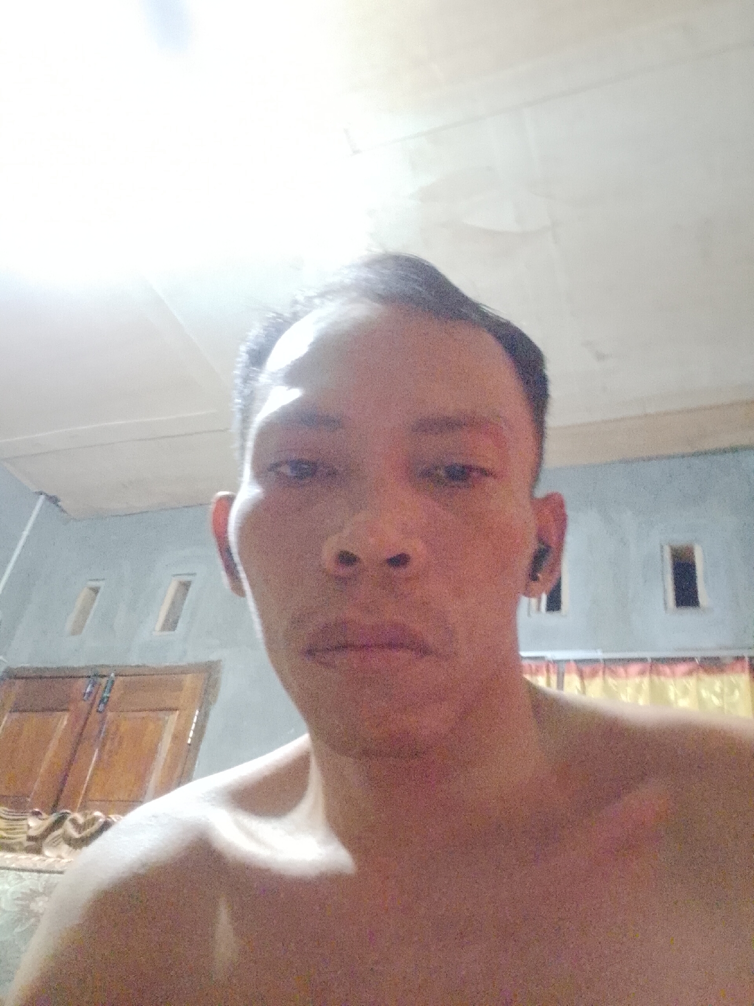 A close-up, front-facing, selfie-style photograph shows a bare-chested individual with short, dark hair. The person is looking directly at the camera. The background features a light-colored ceiling with visible seams, a light blue wall with two rectangular openings, and wooden double doors. The lighting is bright, with a significant glare from above. The image appears to have been taken indoors, possibly in a residential setting in Madukoro, Indonesia.