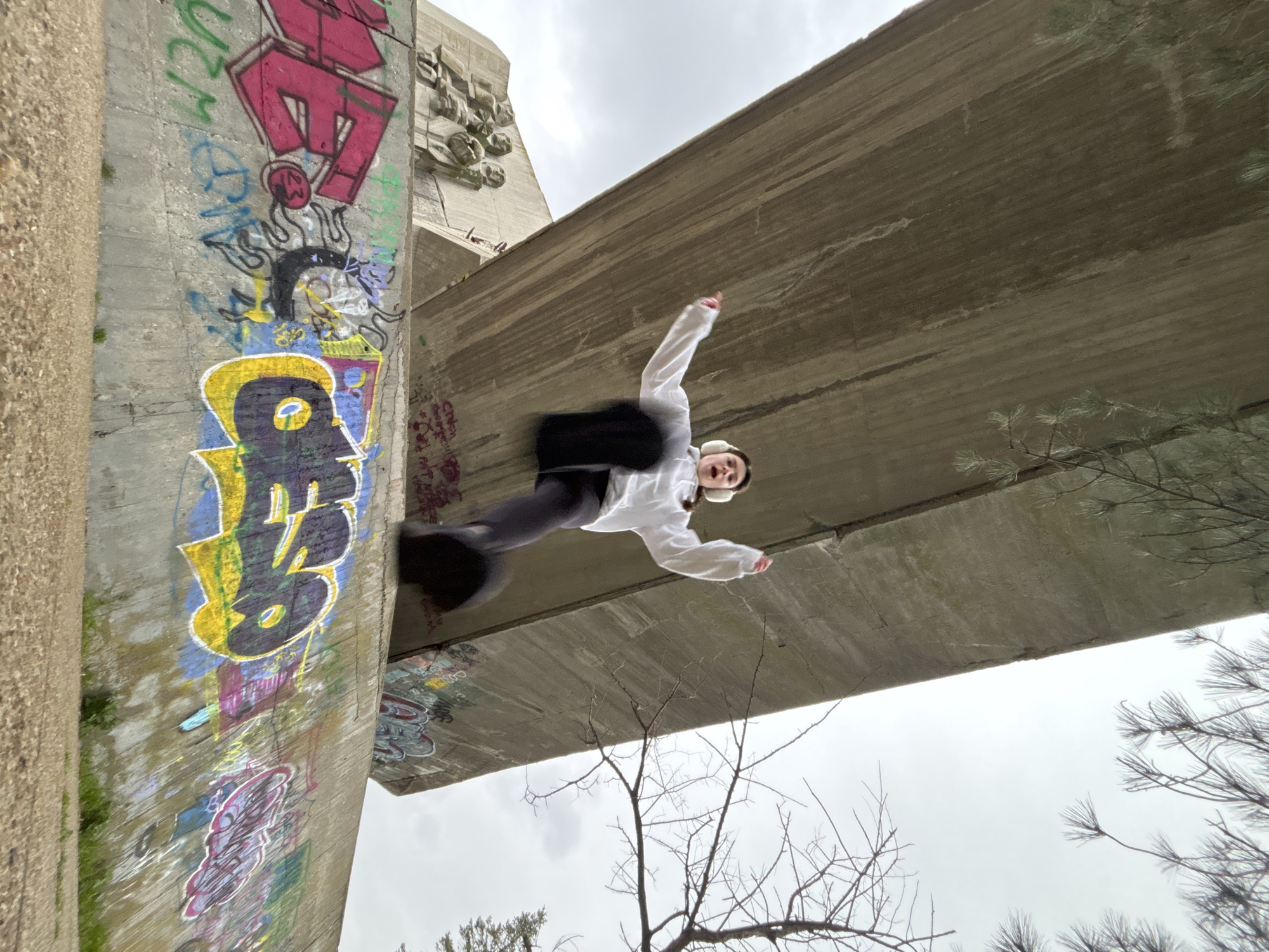 The image is an angled upward shot of a person in motion beneath a concrete structure. The person, a young individual with earmuffs, is wearing a white jacket and dark pants. They appear to be jumping or in mid-air, with arms outstretched. The concrete structure overhead is likely a bridge or overpass, with a rough, textured surface. Graffiti is visible on the vertical support of the structure, featuring a variety of colors and designs. In the background, there are bare tree branches and a gray sky, suggesting a possibly overcast or winter season.
