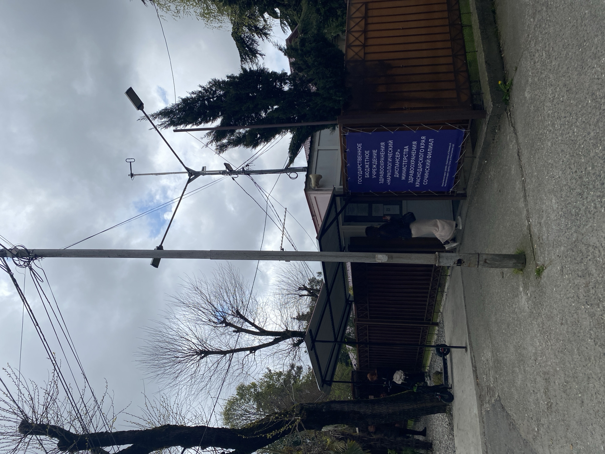 The image shows the exterior of the Sochi branch of the "Narcological Dispensary" of the Ministry of Health of the Krasnodar Territory, Russia. A sign displays this information in Russian. A person in light-colored pants and a dark jacket stands near the entrance to the building. Utility poles with wires are visible in the foreground. Trees, some bare and some evergreen, are nearby. A scooter lies on the sidewalk. The sky is overcast. The address is 354003 Sochi, ul. Plastunskaya, 157.