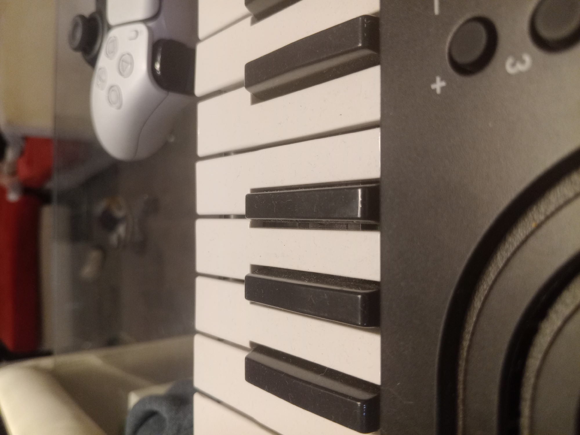 The image shows a close-up view of a keyboard, with a PlayStation 5 controller visible in the blurry background. The keyboard features white and black keys and buttons with markings like "+", "-" and "3". The controller is white. The background is out of focus, showing a sink and other miscellaneous objects. The image location is unknown; there's no information in the provided image that implies Lomas de Zamora, Argentina.