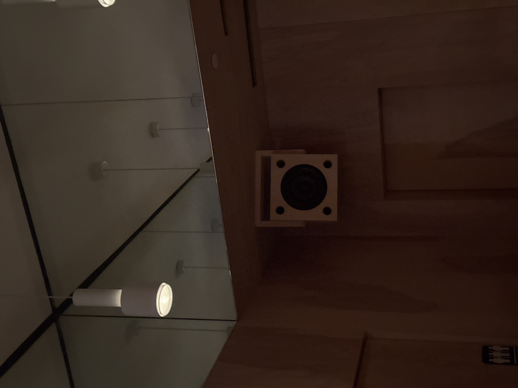 The image shows a close-up interior view. On the left, there's a mirrored surface reflecting a series of circular lights. To the right of the mirror is a wooden surface. A square speaker with a black center and four visible screw holes is mounted on the wooden surface. Below the speaker, at the bottom-right corner, is a sign with a male and female figure, which often indicates restroom facilities. The setting appears to be indoors, possibly a sauna. The location is unknown.