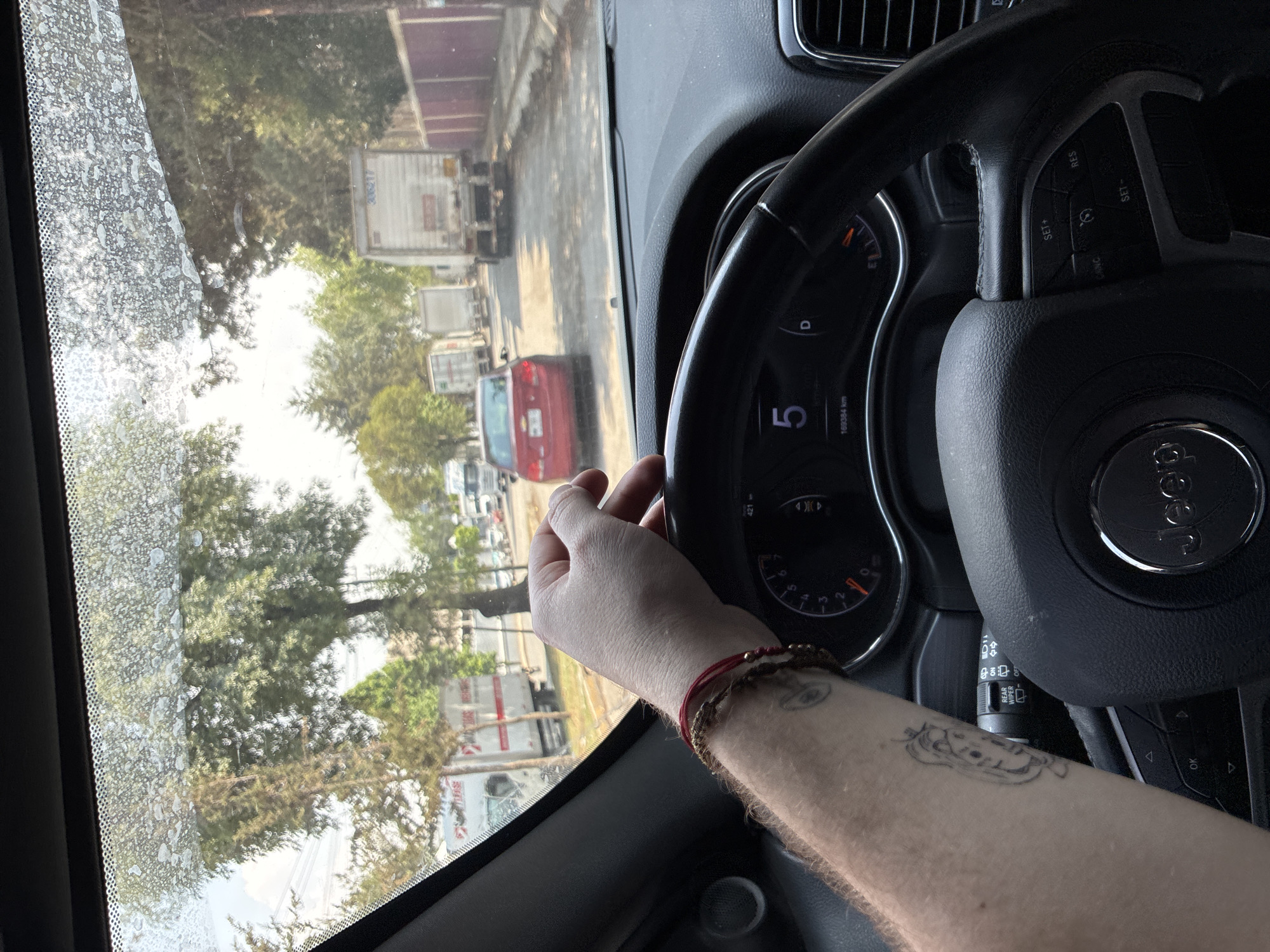 The image shows the interior of a vehicle, specifically a Jeep, from the driver's perspective. The steering wheel is in view with a "Jeep" logo on the center. A hand is partially visible, resting on the steering wheel, with bracelets and a tattoo on the forearm. The dashboard is partially visible, displaying the speedometer reading 5 and the odometer reading 169384 km. Through the dirty windshield, the outside view shows a street with various vehicles, including a red car and several white delivery trucks or trailers. The environment suggests a suburban or industrial area with trees lining the roadside. Based on the OCR of "36217" and location information, the city is Cuautitlán Izcalli, Mexico.