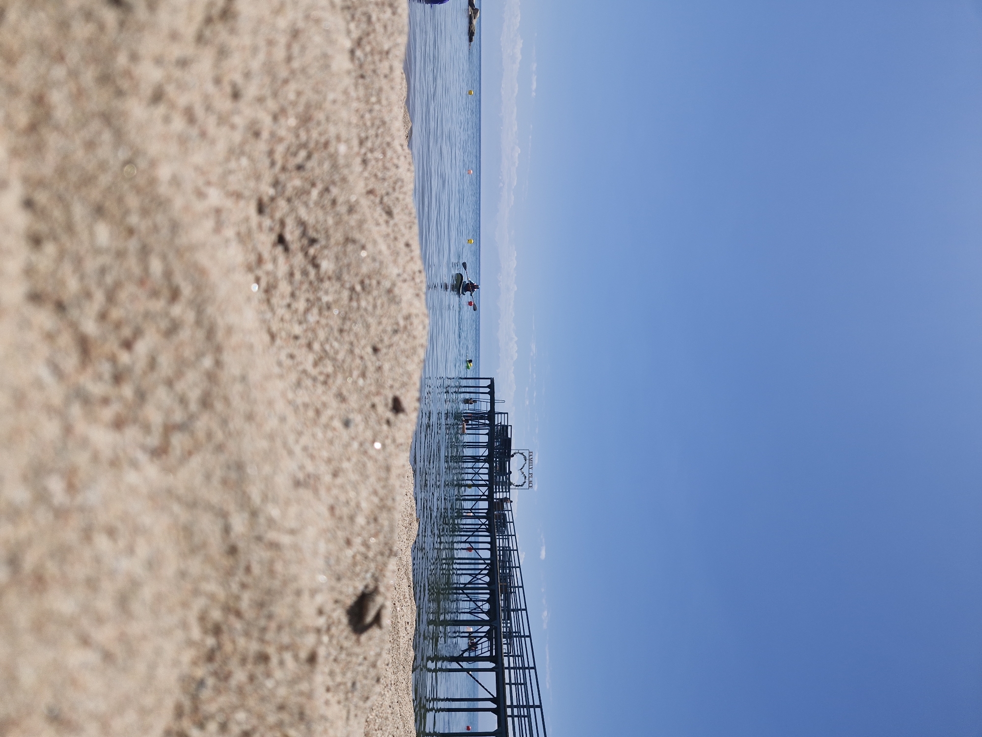 A low-angle, long shot captures a sandy beach in the foreground with the calm, blue water of Lake Issyk-Kul and a wooden pier extending into the distance under a clear, bright blue sky. The pier has a railing and supports, and a sign that reads "FAMILY PLACE" with a heart shape is visible on it. Several people are present on the pier and in the water. One person is kayaking in the middle distance. Bouyant markers are spread across the water. In the far distance, hazy mountains are visible along the horizon. The scene appears to be a recreational area on the north shore of Lake Issyk-Kul, near Bulan-Segestu, Kyrgyzstan, during daylight hours.