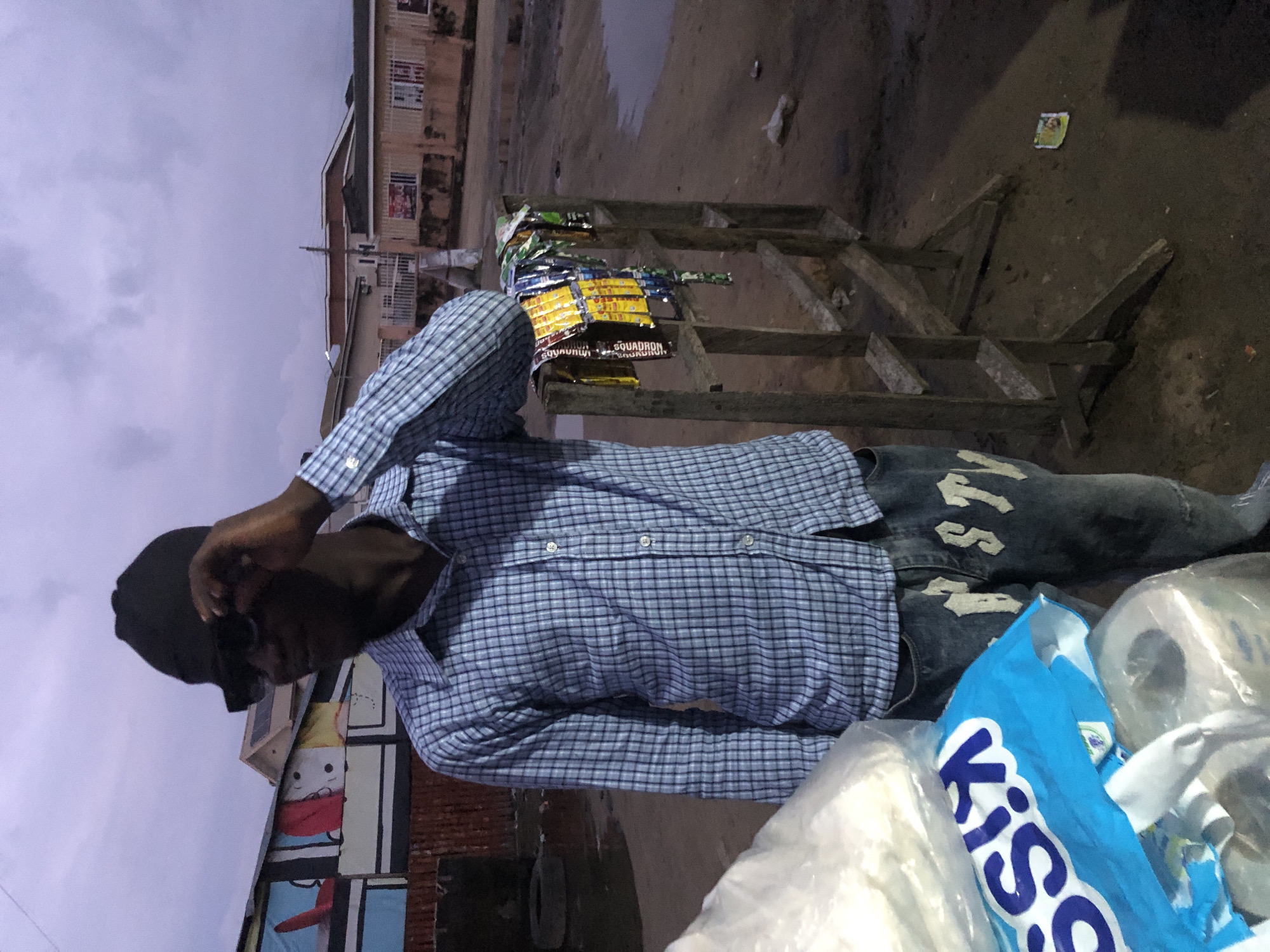 A young Black man wearing a blue plaid shirt, black beanie, and distressed jeans stands in a city street. He is adjusting his dark sunglasses with his right hand. The man is positioned in front of a makeshift wooden display rack holding various colorful snack packets. Behind him, buildings with painted murals and other structures are visible. The ground is dirt and appears wet in places, suggesting it may have rained recently. The sky is overcast and a muted purple-gray color. The scene is consistent with a street vendor operating in Ado, Nigeria, likely in a market or busy thoroughfare. In the foreground to the left are several plastic-wrapped items, including a blue bag with the word "KISS" printed in white.