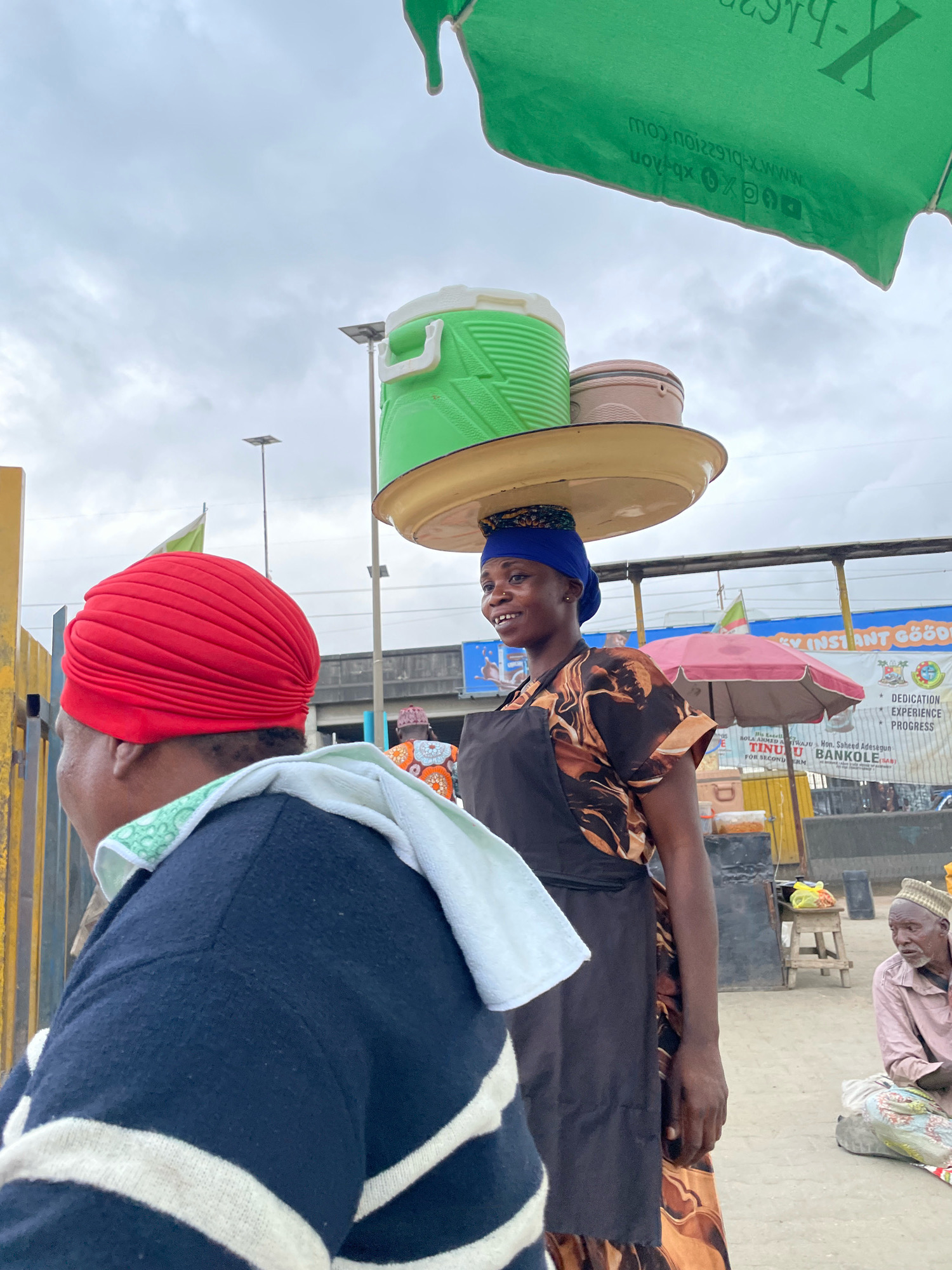 A woman with a blue headscarf is carrying large green and tan containers balanced on a yellow platter on her head. She wears a brown and black patterned dress and a dark apron. She is standing outdoors, with a large green canopy overhead. In the foreground, a person wearing a red headscarf and a dark sweater with white stripes is partially visible. In the background, another person, an elderly man, is seated on the ground. The setting appears to be a market or street scene in Ajah, Nigeria, indicated by signage visible in the background that includes "BANKOLE" and other text in English and possibly a local language. The environment suggests an overcast day.