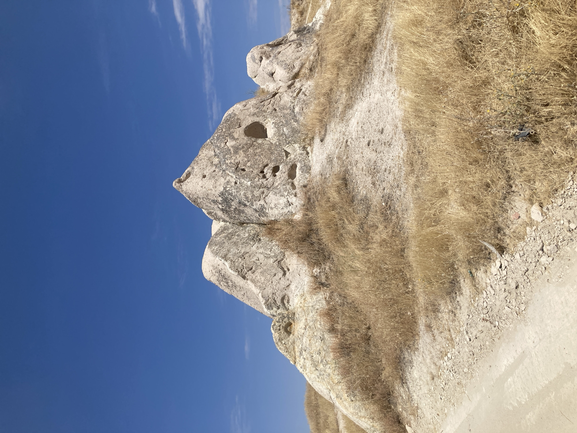 A scenic view of the Cappadocian landscape, specifically the unique rock formations characteristic of Göreme, Turkey. The image features a cluster of light-colored, weathered rock formations that appear to have been sculpted by natural erosion. One prominent formation in the center has a pointed peak and several openings or cave-like structures carved into its surface, suggesting historical human activity. The rocks are situated on a gentle slope covered in dry, golden-yellow grass and sparse vegetation. In the foreground, a dirt path or road curves into the scene from the lower left, with loose gravel and small stones along its edge. The background is dominated by a clear, vibrant blue sky with a few wispy white clouds. The overall impression is one of a dry, arid environment with striking geological features.