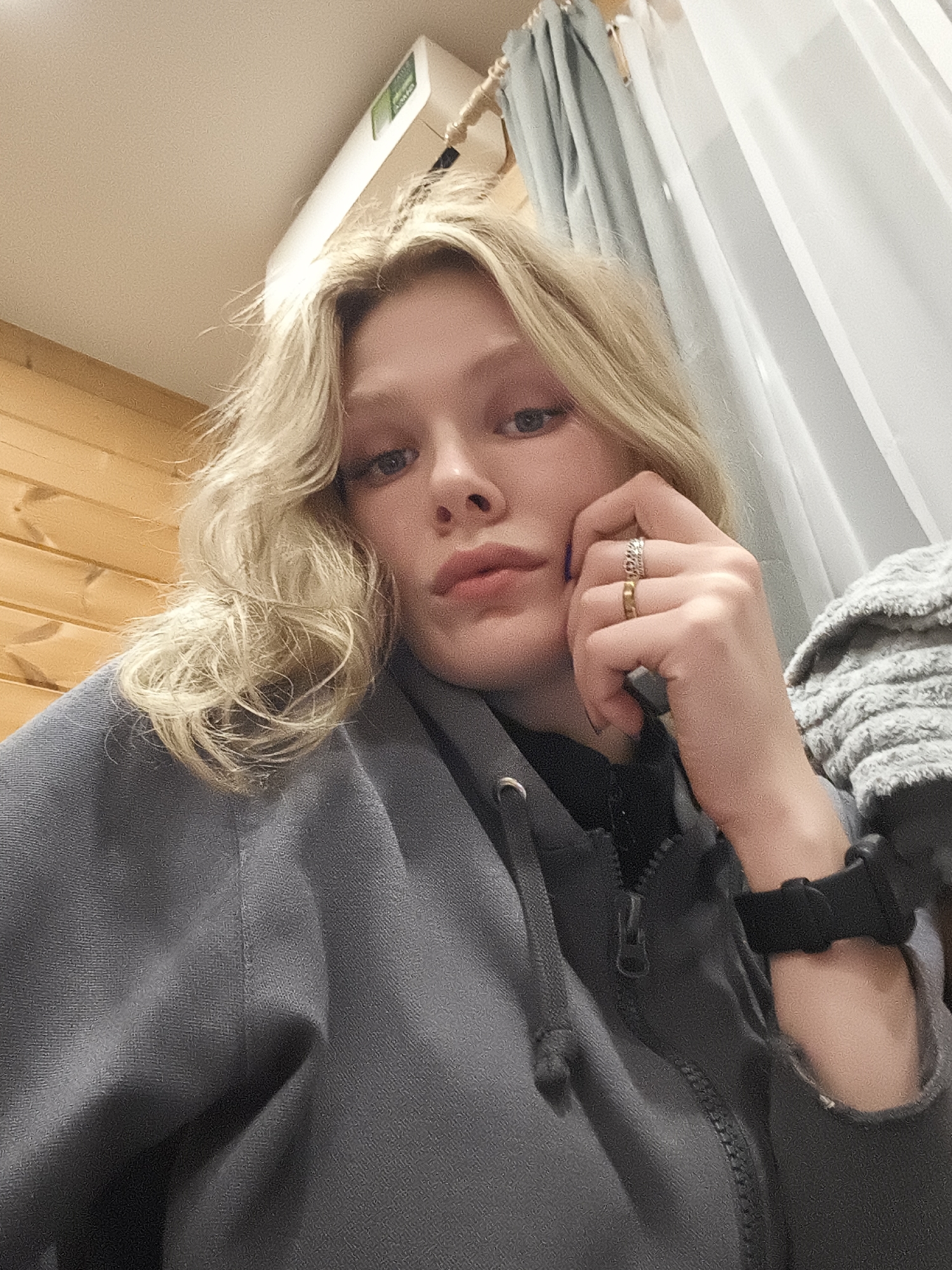 A selfie depicts a young woman with blonde, wavy hair and fair skin. She is wearing a gray hooded sweatshirt with a zipper partially visible. Her left hand is raised to her face, with two rings visible on her fingers. A black wristband is on her left wrist. Behind her is a wooden wall, a white ceiling, and light blue curtains with a white sheer curtain behind them. The location is Русский Вожой, Russia.