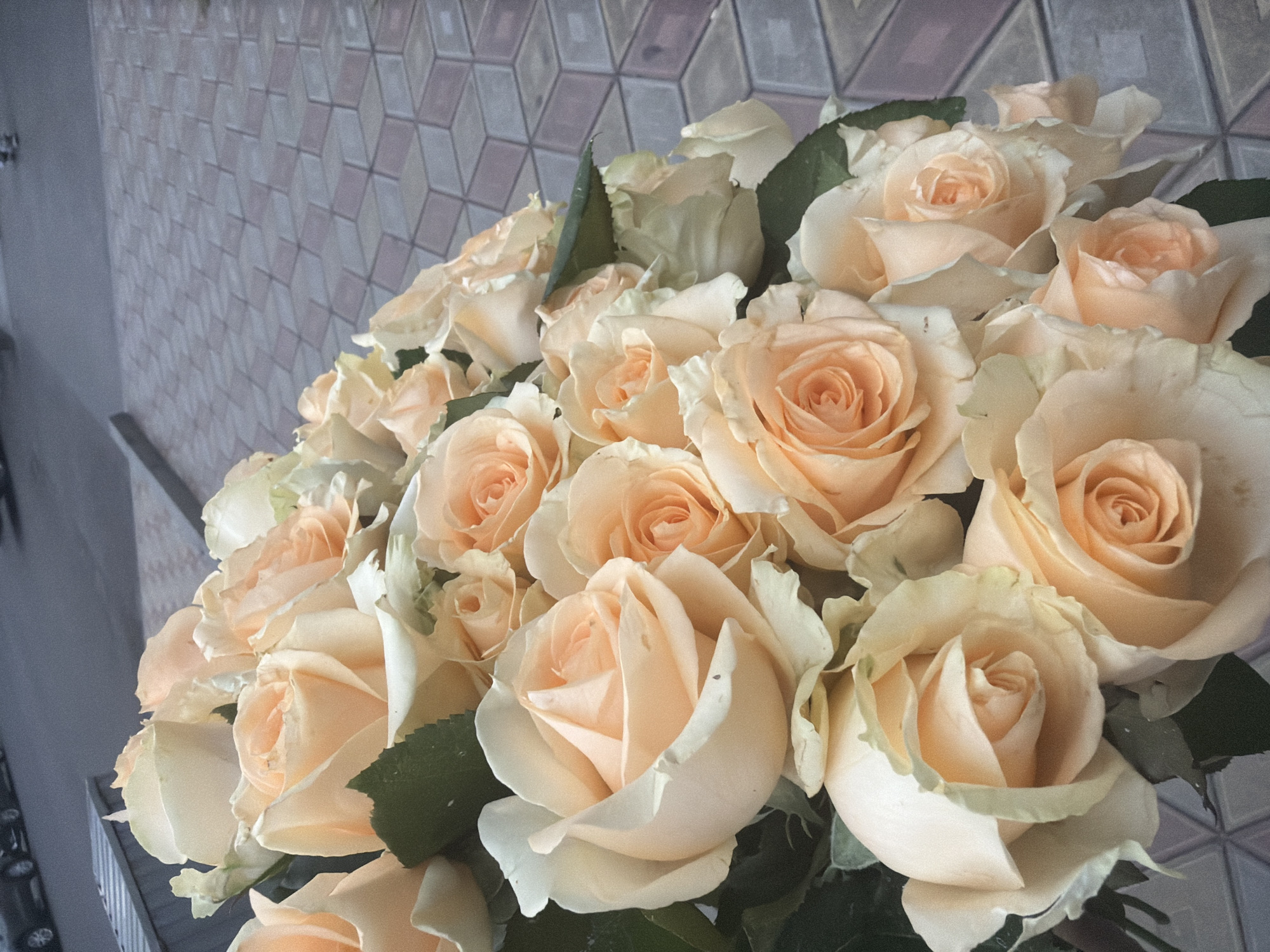 A bouquet of light peach and cream colored roses is displayed outdoors on a patterned paved surface. The paving stones are laid in a geometric design of interlocking diamond shapes in shades of grey and pink. In the upper left quadrant of the image, a dark grey paved surface is visible, possibly a street or sidewalk, with the tires of a vehicle and some blurry figures in the background, suggesting an urban setting. The roses, some fully bloomed and others partially open, fill the majority of the frame, with their soft petals and green leaves. The lighting appears natural, casting subtle shadows on the petals and the pavement. The overall impression is one of a floral arrangement placed outdoors in an urban environment.