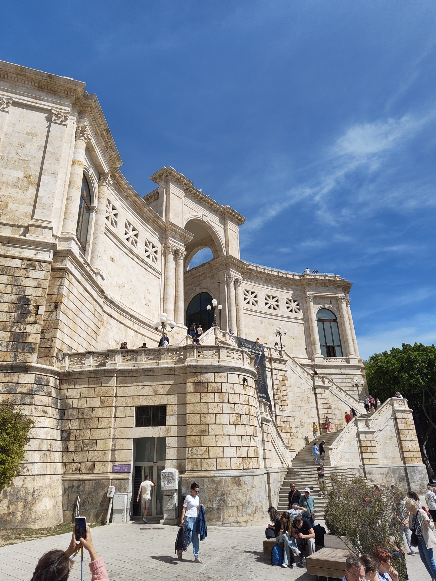 The image shows the Bastione di Saint Remy, a large white stone monument in Cagliari, Italy, under a partly cloudy blue sky. The architecture includes arches, columns, and decorative windows. A large banner hangs from one section of the building. People are present in the scene: some are sitting on benches in the foreground, some are climbing the stairs leading up to the monument, and others are walking near the entrance. A woman in the foreground is taking a photo with her phone. A tree is visible on the right side of the image.