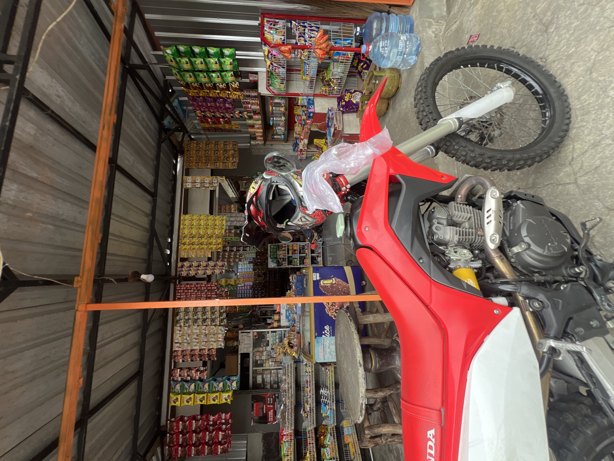 The image depicts a red and white dirt bike parked in front of a small store, likely a warung, in Rendang, Indonesia. The warung is open-air, with a corrugated metal roof supported by orange and black metal beams. Shelves are laden with various packaged snacks and drinks, with some items hanging from the ceiling. A glass-fronted freezer unit displays ice cream. In the foreground, the dirt bike's red seat and white fairing are prominent. A helmet, covered partially in plastic wrap, rests on the handlebars. Behind the bike, a rough-hewn wooden table and stools are visible. Two large plastic water jugs are stacked near the entrance of the store. The ground is paved concrete. The overall scene suggests a typical roadside convenience store in a rural or semi-urban Indonesian setting.