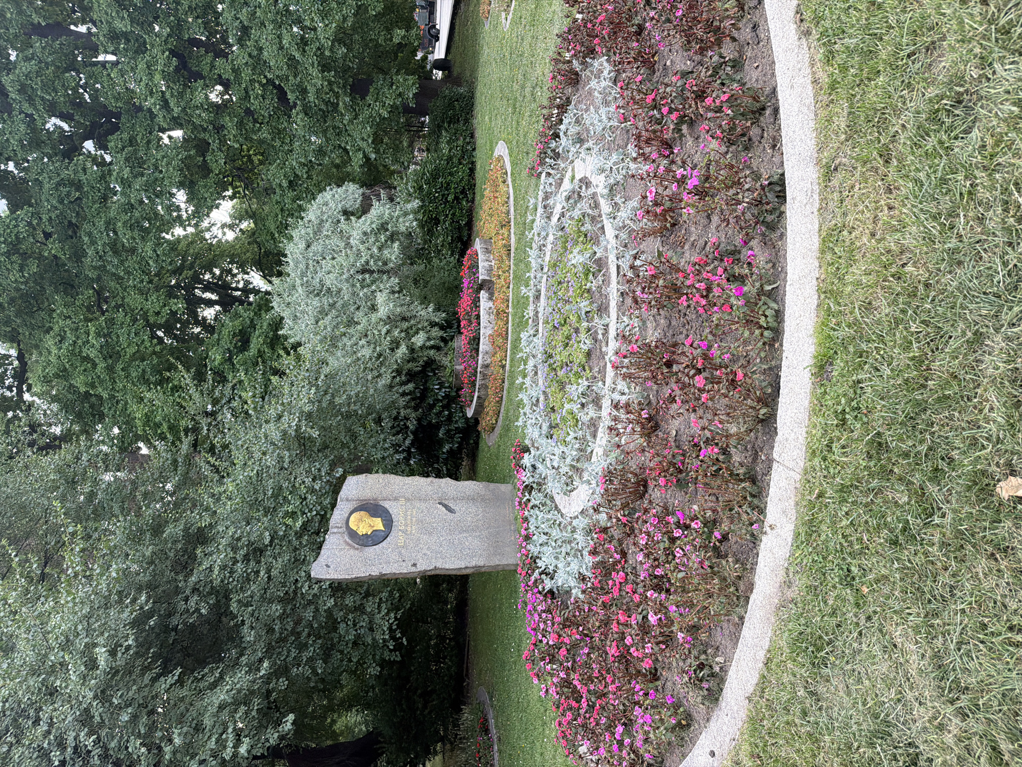 A memorial is situated in a green park in Sofia, Bulgaria. A tall, rough-hewn granite stone monument stands in the background, featuring a round golden medallion with a profile of a person's head. Below the medallion, text is inscribed in Cyrillic: "ЦАР БОРИС III" and dates "1894 – 1944". In front of the monument, a circular flower bed is planted with various flowers, including pink and deep red blooms, and silvery-green foliage plants arranged in concentric rings. Another, smaller flower bed with red and orange flowers is visible further back. The ground is covered in lush green grass, and large trees with dense foliage dominate the background, indicating a park setting within the city. The lighting suggests it is daytime, possibly overcast, as there are no harsh shadows.