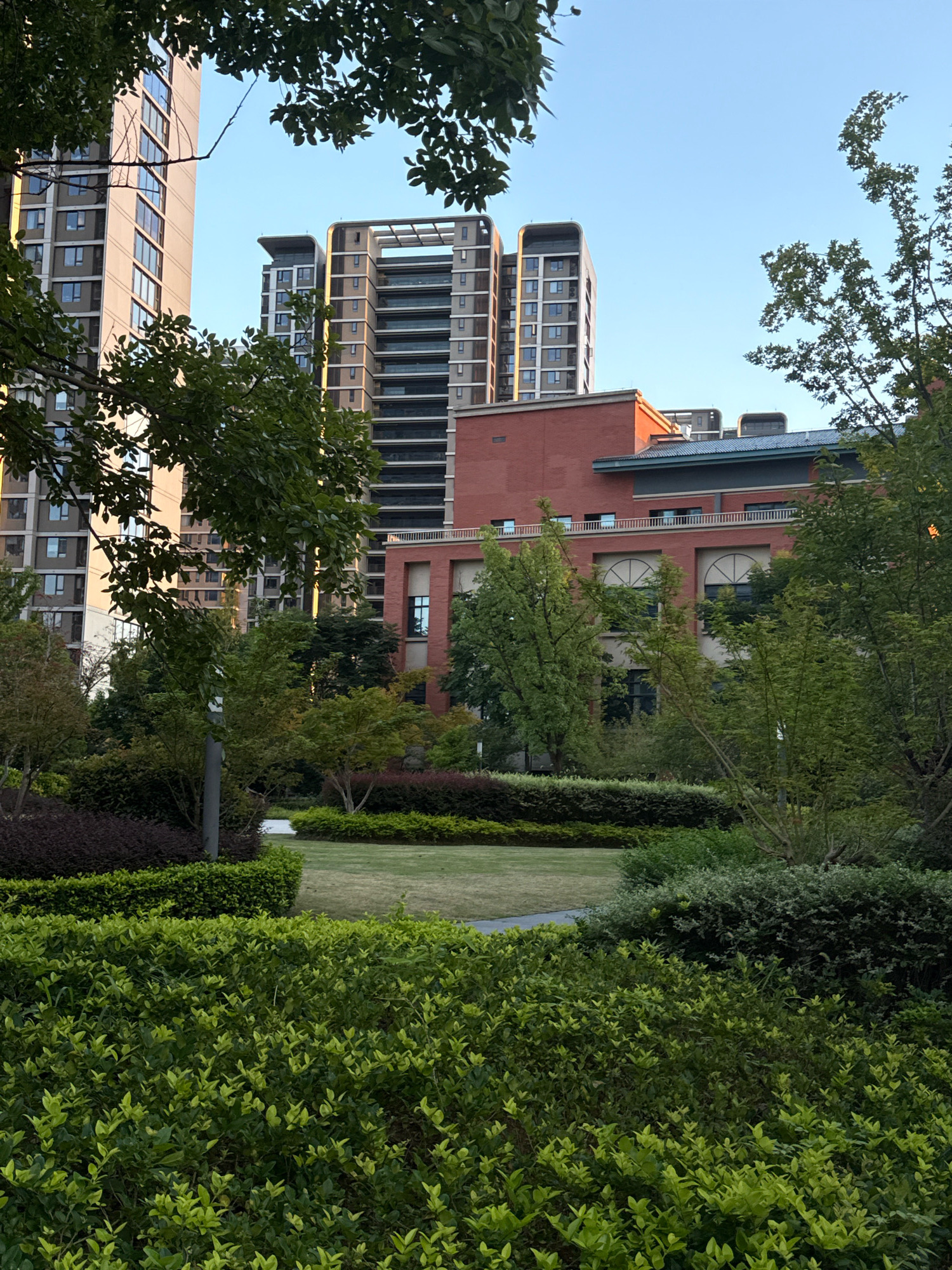 The image depicts a landscaped area in the city of Yiwu, China, with modern high-rise buildings visible in the background. In the foreground, lush green bushes and manicured hedges form a dense layer, with a pathway and a patch of grass visible further back. Behind the greenery, a red brick building with arched windows and a white railing on a balcony is present. Beyond that, several tall, contemporary residential buildings with numerous windows rise into the clear blue sky. The scene appears to be a well-maintained urban park or communal space, likely a residential complex. There are no people or animals visible in the image, and the overall atmosphere is calm and serene, bathed in what appears to be late afternoon sunlight.