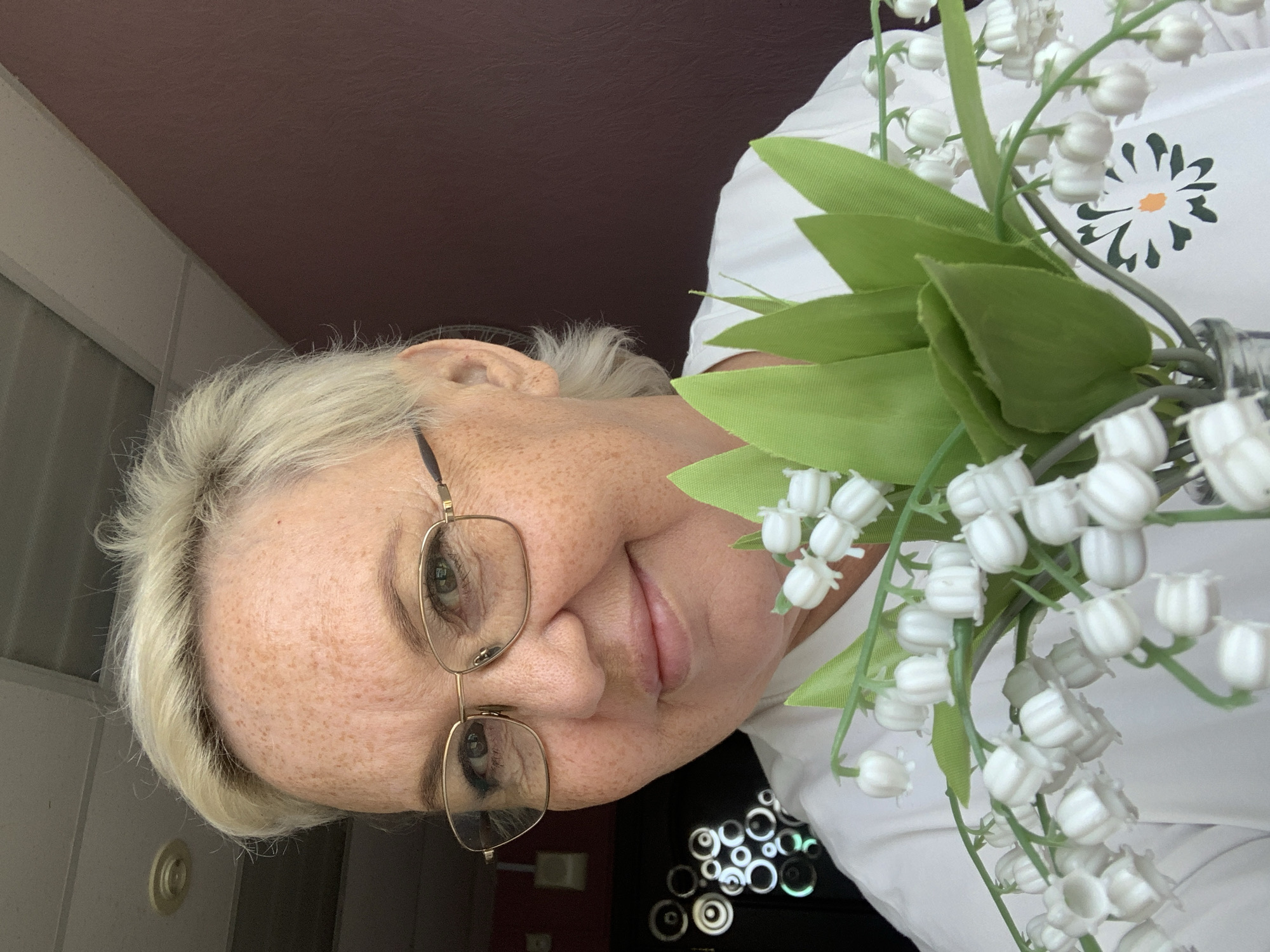 A woman with short blonde hair and glasses holds a bouquet of artificial lily of the valley flowers. She is wearing a white t-shirt with a daisy graphic. The background consists of a wall, a ceiling, and a patterned door. The scene appears to be indoors, possibly in a residential setting. The image was taken in Chaikovsky, Russia.