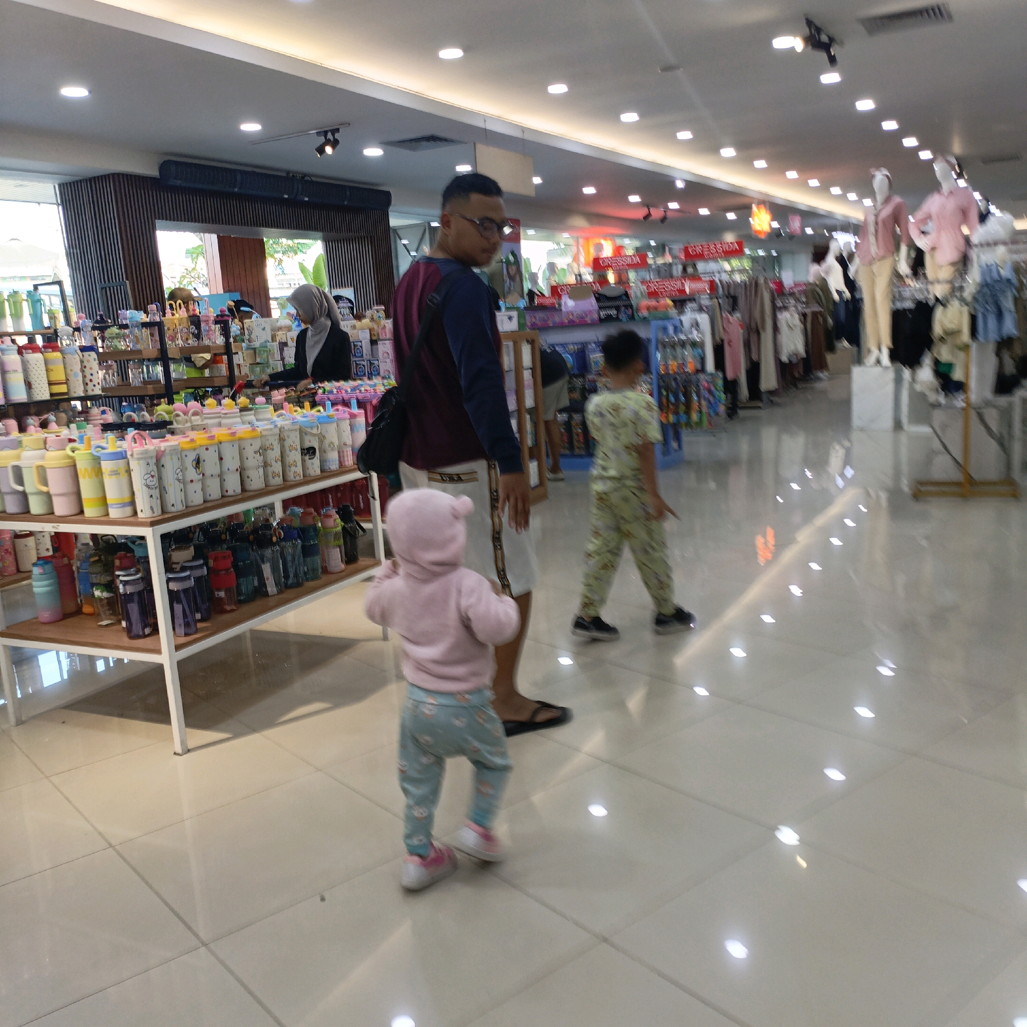 The image depicts an interior scene of a store, likely a department store or a large retail space, in Salatiga, Indonesia. The floor is a light-colored tile, highly reflective, with numerous ceiling lights casting bright, square-shaped reflections. In the foreground, a toddler wearing a pink hooded jacket and blue patterned leggings with pink and white shoes is walking away from the viewer. To the left and slightly behind the toddler is an adult male wearing glasses, a dark long-sleeved shirt with a maroon yoke, and white shorts. He is looking to his right. Further in the background, another child, a boy, is walking to the right, wearing a light green patterned outfit. The store displays various merchandise on shelves and racks. Notably, a shelf in the mid-left foreground is filled with numerous colorful water bottles. In the background, clothing racks with garments and mannequins are visible, suggesting a section selling apparel. The overall lighting is bright and even, typical of a retail environment. The scene appears to be a casual outing for the individuals pictured.