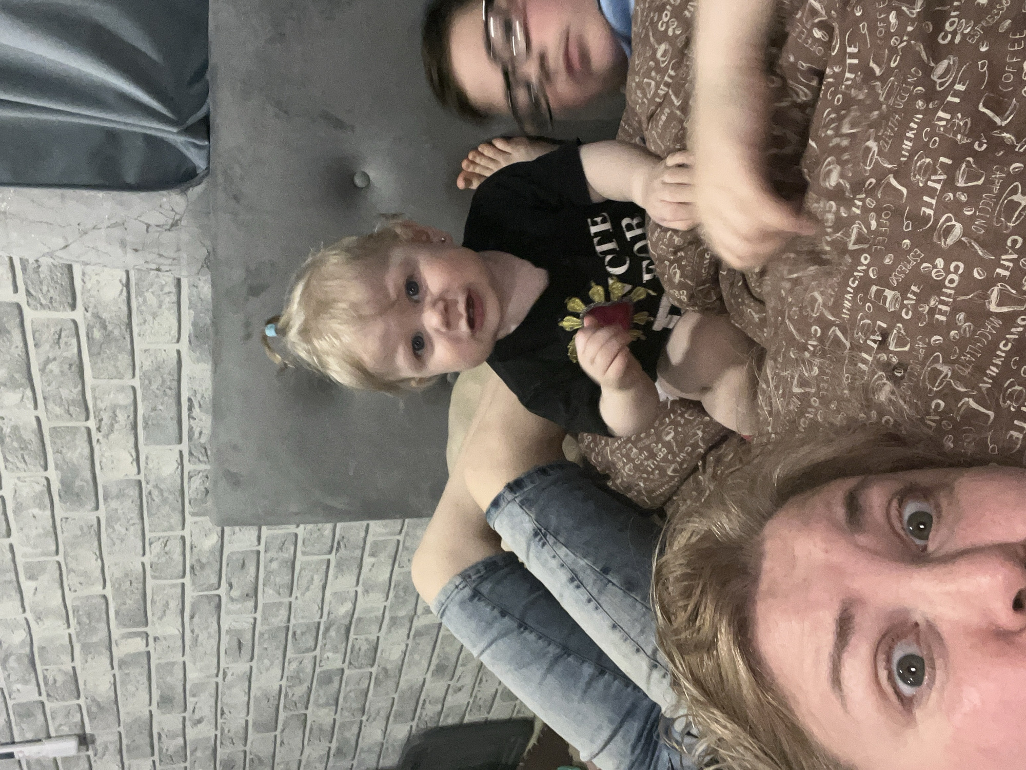 A selfie is taken from a low angle capturing a woman, a child, and another person lying on a bed. The woman, positioned at the bottom of the frame, has wide eyes and fair hair. She is wearing blue jeans. Adjacent to her is a young child with fair hair pulled back in a ponytail, wearing a black t-shirt with a sunflower design and the word "PROTECT" printed on it. Behind the child, there is another individual wearing glasses. All three individuals are lying on a brown patterned duvet with coffee-related graphics printed on it. The background includes a grey headboard and a wall with a brick-like pattern. The photo was taken inside an interior, most likely a home. The exact location in Vesyoloye, Russia is not identifiable from the image.