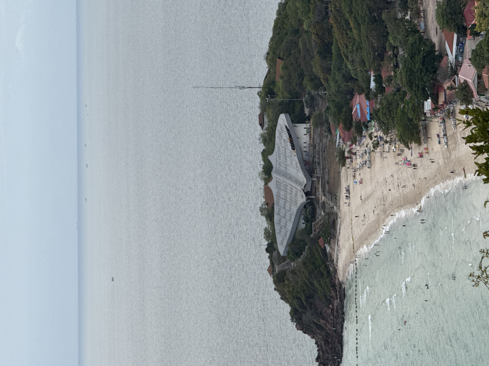 The image depicts a coastal scene in Pattaya City, Thailand. A beach with light-colored sand is populated with numerous people engaging in recreational activities. Adjacent to the beach, a rocky headland rises, covered with dense vegetation and structures including buildings and an architecturally distinctive, modern building with a white, angular roof. A tall communications tower is situated on the headland. The water appears calm with gentle waves lapping the shore. In the background, the sea extends to the horizon under a cloudy sky.