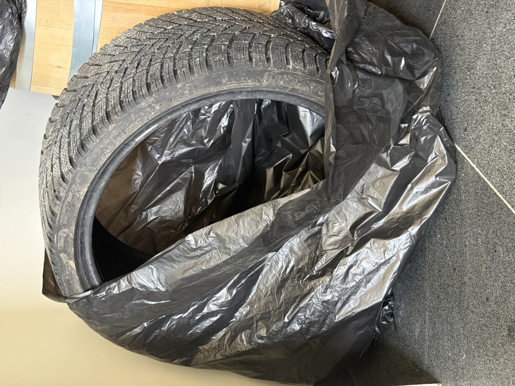 A winter tire is partially concealed within a crumpled black plastic bag. The tire rests on a speckled dark gray floor, likely a paved surface. In the background, a portion of a light-colored wall and a section of a wooden barrel are visible. The overall environment appears to be indoors, possibly in a storage area or garage, rather than an outdoor urban setting in Dolno Ablanovo, Bulgaria. There are no people or any indication of human activity other than the placement of the tire.