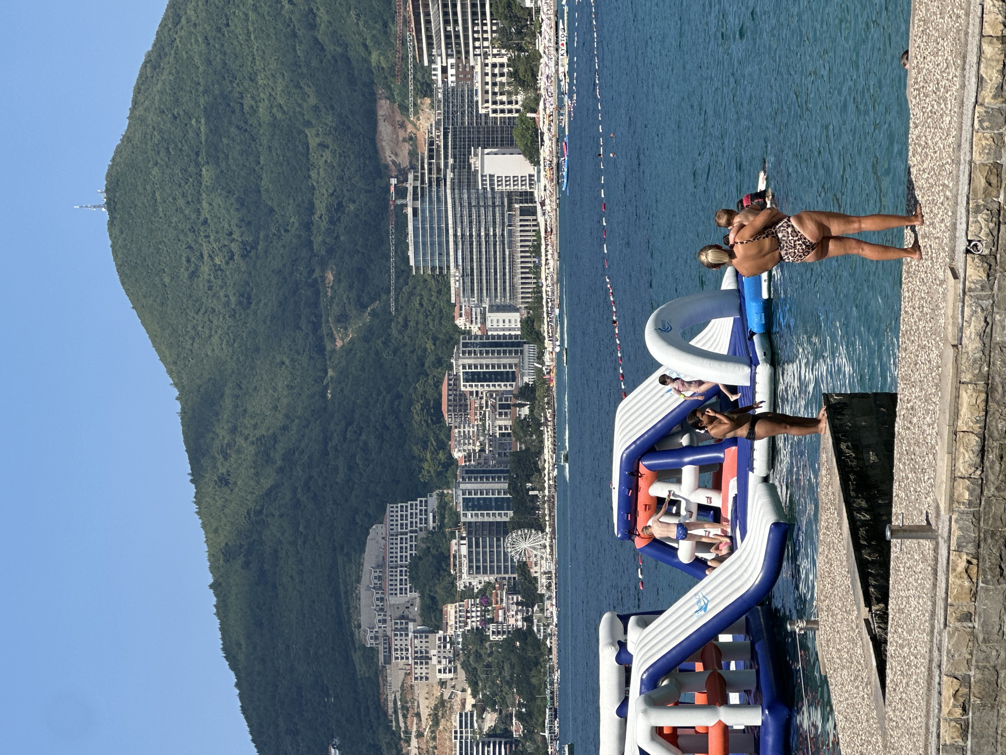 The image depicts a sunny day in Boreti, Montenegro, with a view of the Adriatic Sea and a backdrop of a verdant, mountainous landscape. In the foreground, two inflatable water park structures are partially submerged in the blue water. On a stone walkway in the lower right of the frame, a woman in a leopard-print swimsuit holds a baby, facing the water. To her left, a young child in blue swim trunks stands on one of the inflatable structures, looking out. Another child in a pink swimsuit is visible on the same inflatable. In the middle ground, the coastline is lined with modern buildings and hotels, with a Ferris wheel visible among them. The background is dominated by a large, tree-covered mountain with a communication tower on its summit. The water is calm, with several buoys and ropes delineating areas of the sea.